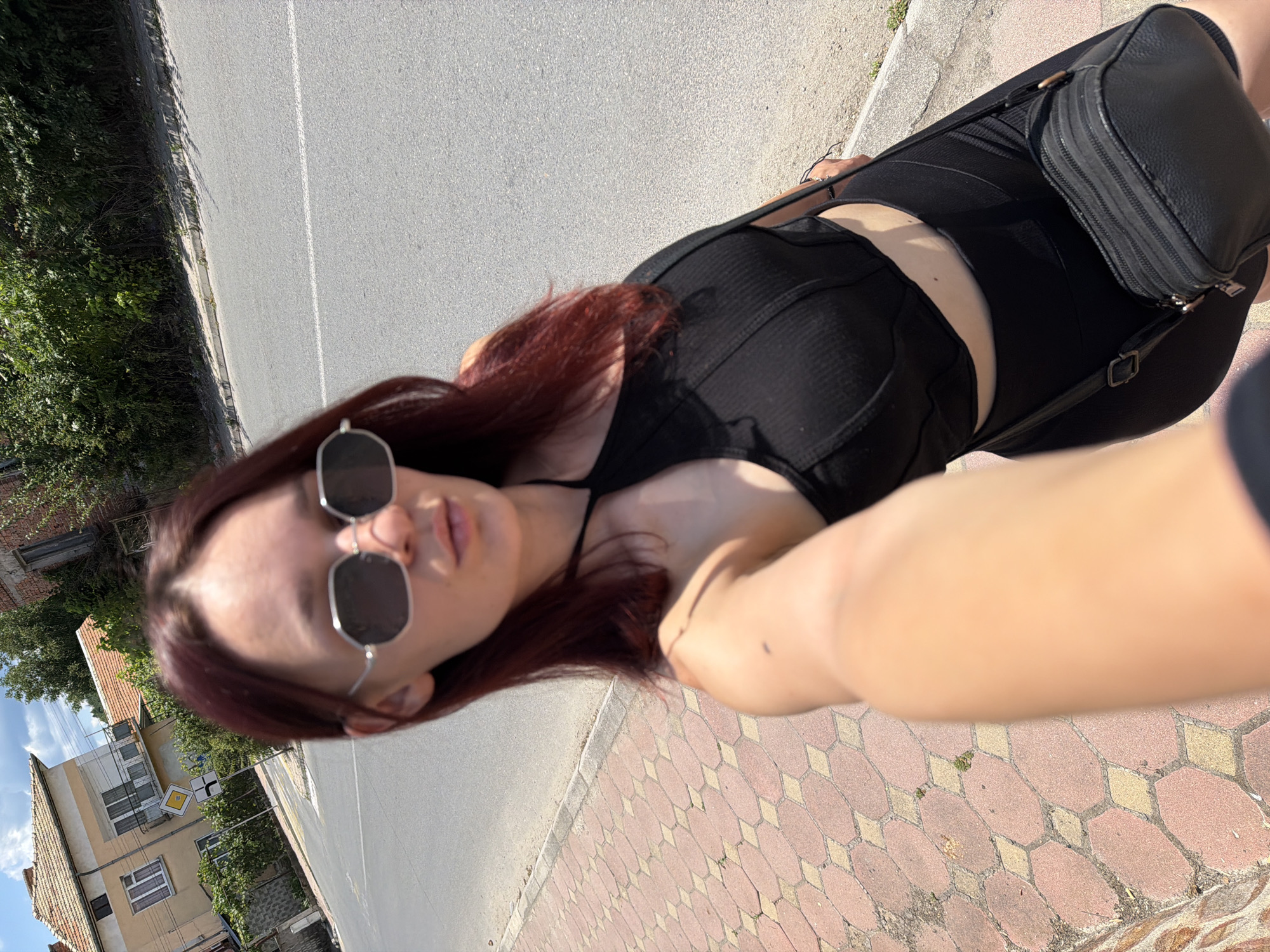 A young woman with reddish-brown hair is taking a selfie outdoors in what appears to be a village or small town. She is wearing a black sleeveless top and black shorts, and a black crossbody bag is slung over her shoulder. She has on large, hexagonal-shaped sunglasses. The background shows a street with a sidewalk made of red and yellow patterned paving stones. To the left of the street, there is a dense green bush and a brick building with an orange-tiled roof. Further in the background, more buildings with similar roofs and trees are visible. A yellow diamond-shaped road sign with black arrows is also visible on a post near the road. The lighting suggests it is daytime, possibly late morning or afternoon, with bright sunlight casting shadows. The overall scene appears casual and relaxed, with the woman looking directly at the camera with a neutral expression. The location context indicates this is Hadzhidimovo, Bulgaria. No visible text is present in the image.