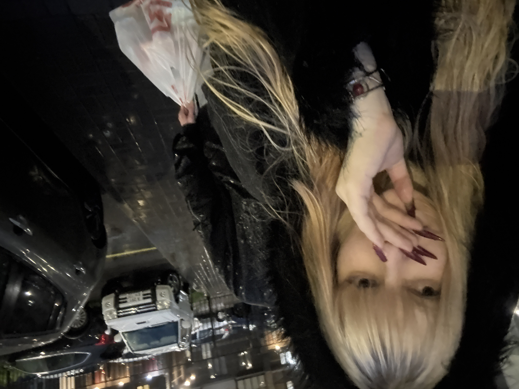 A young woman with blonde hair and long fingernails stands outside at night, holding a white plastic bag in her right hand. She is wearing a black coat with a fur-lined hood. In the background, several cars are parked on a wet street. The scene appears to be in an urban setting, possibly a residential area, given the presence of parked cars and buildings. The lighting suggests it is dark, with some artificial lights from the buildings visible.