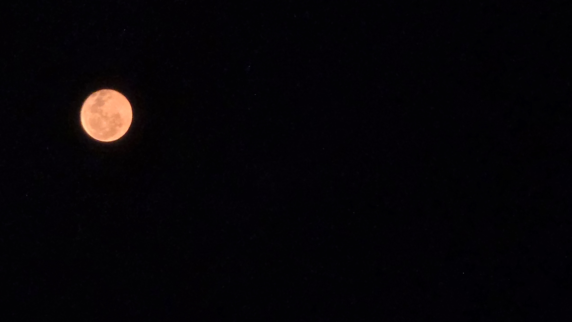 The image displays a full moon against a dark, star-speckled sky. The moon has an orange hue and is positioned in the upper right quadrant of the frame. The background is entirely black, suggesting it is nighttime. There are no other discernible objects or features in the image. The location of Velykyi Zhytyn, Ukraine, is not evident from the visual information presented in the image.