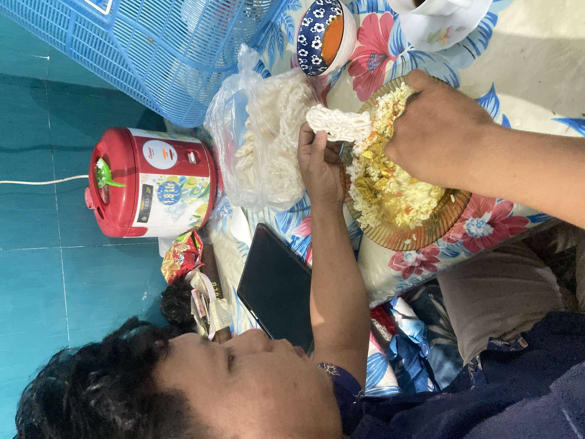 A person is eating rice with a curry and is holding a piece of white crackers above the plate. The person is wearing a blue shirt and is seated at a table covered with a floral tablecloth. A red rice cooker, a blue plastic basket, and a bowl of orange sauce are visible on the table. The background appears to be a wall with blue tiles. The scene is indoors, likely in a residential setting in Gelam, Indonesia.