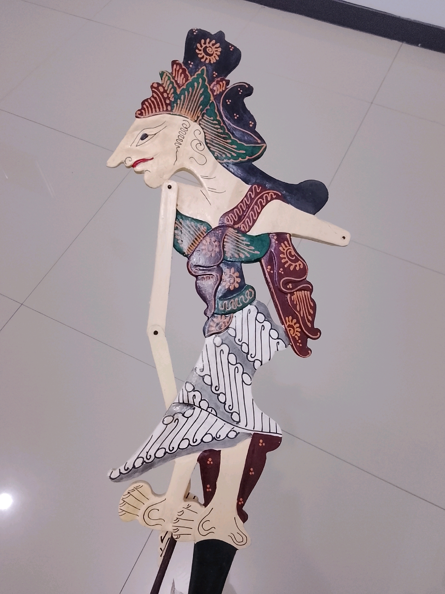An ornate, traditional Indonesian Wayang Golek puppet, likely made of wood and hand-painted, stands upright on a light gray, tiled floor. The puppet is depicted in profile, facing left, with a pointed nose, red lips, and a detailed headdress featuring dark blue, green, and red elements. It wears a decorative garment with floral and patterned motifs. The puppet has visible wooden joints and is supported by a dark base. The background consists of the tiled floor, with a dark, angled line visible in the upper right quadrant, possibly a shadow or an architectural feature. A bright, circular light reflection is present on the floor in the lower left. The scene suggests the puppet is either on display or being prepared for a performance, possibly within a cultural institution or venue in Slawi, Indonesia, a region known for its traditional arts.