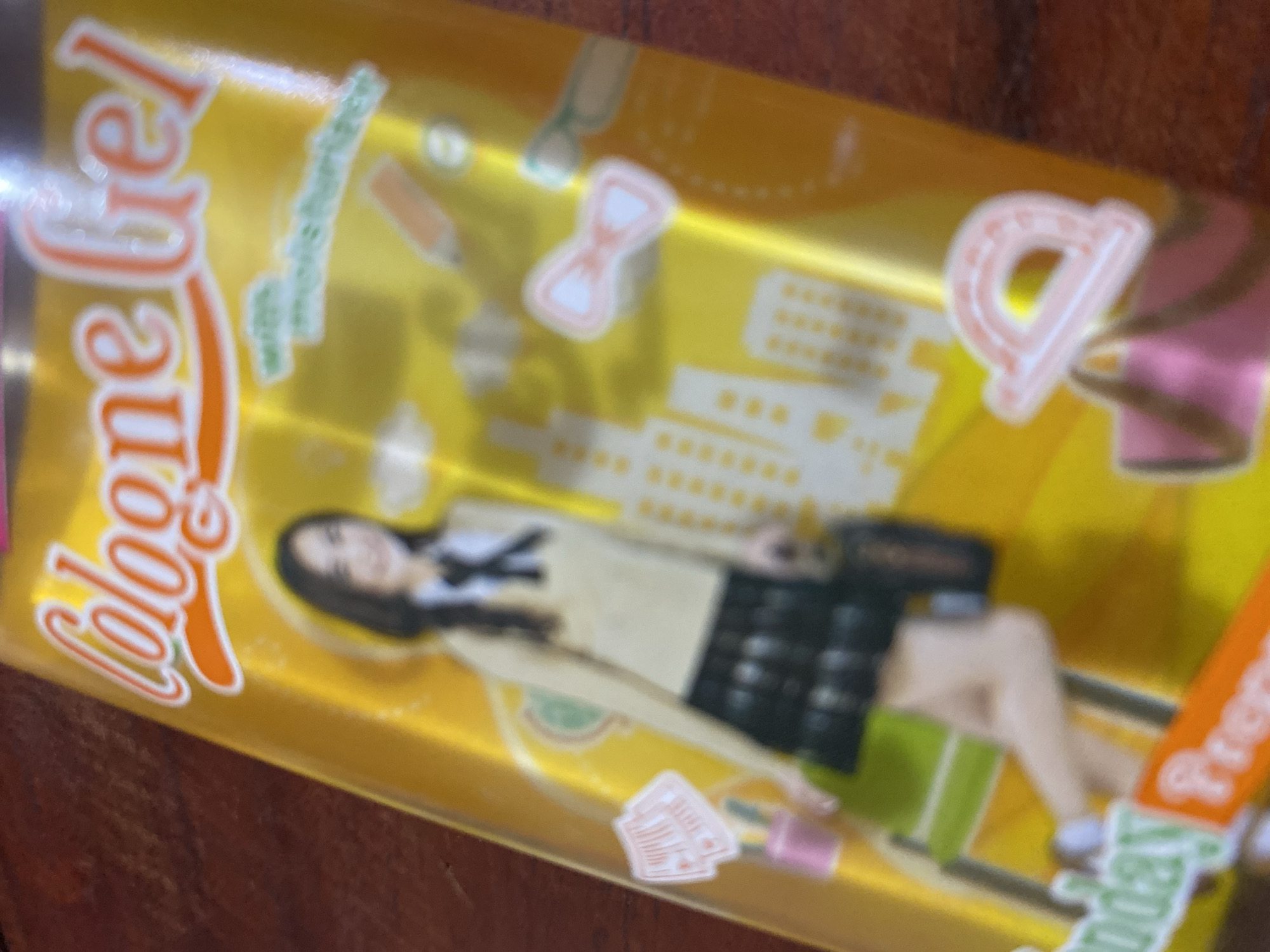 The image shows a close-up of a product with a yellow background. The product appears to be a gel or lotion, with the text "Cologne Gel" prominently displayed in orange and white lettering at the top. Below the text, there is an illustration of a young woman in a school uniform, consisting of a beige sweater, white shirt, and plaid skirt. She is holding a black bag. Scattered around the illustration are various school-related icons, such as a pencil, a bow, and a protractor. The overall aesthetic is bright and playful, suggesting it is marketed towards a younger demographic. The background is blurry, but it seems to be a textured surface, possibly wood. There is no information in the image that indicates a specific location in Ta', Indonesia.