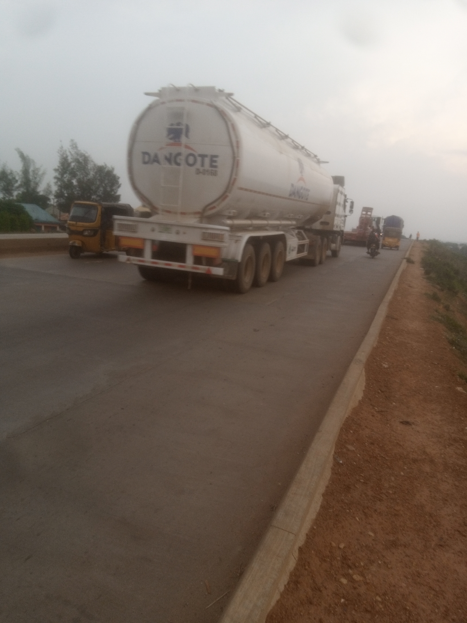A long white tanker truck with "DANGOTE" written on its side is parked on a paved road. The truck has multiple axles and a large cylindrical tank. To the left of the tanker truck, a yellow three-wheeled vehicle is visible. In the background, there are trees and structures, suggesting an outdoor setting, possibly in a semi-urban or rural area. The sky is overcast and hazy. Further down the road, a motorcycle and another truck are present. The road itself appears to be in good condition, with a dirt shoulder on the right side. The overall lighting suggests it might be late afternoon or early morning.