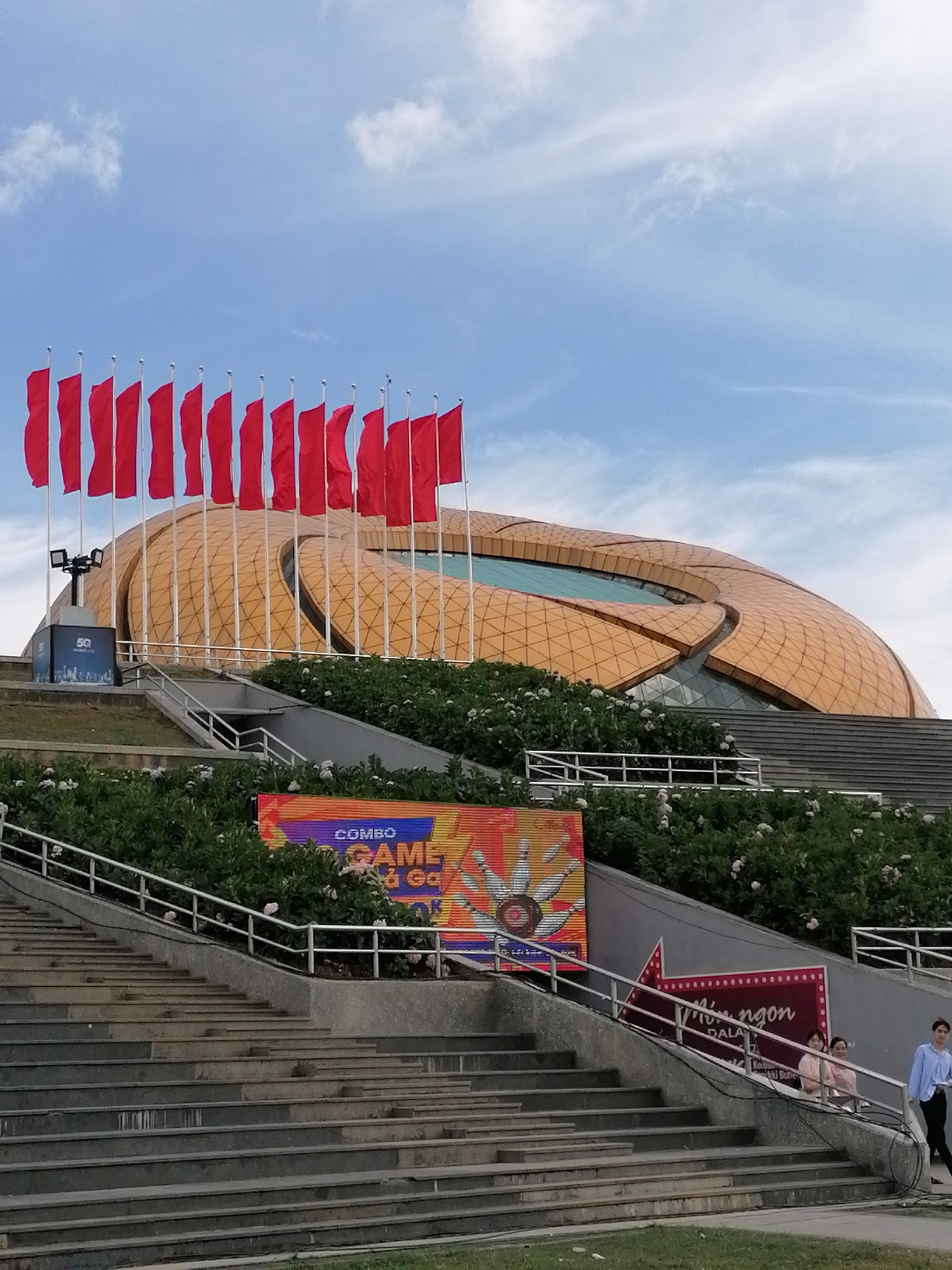 In Da Lat, Vietnam, a grand, golden-domed building with a distinctive geometric design is visible under a blue sky with scattered clouds. A series of red flags on poles line the foreground, leading towards the building. In front of the building, a large set of concrete stairs descends, bordered by lush green bushes and white flowers. At the bottom of the stairs, a vibrant, colorful digital billboard displays the words "COMBO GAME" and imagery of bowling pins. Further down the stairs, another illuminated sign, shaped like a red arrow with white dots, features Vietnamese text. Two women stand on a landing near the arrow sign, observing the scene, while a young man in a blue shirt walks down the stairs on the right. The overall impression is of a public gathering space or entertainment venue, likely in a park or plaza setting.
