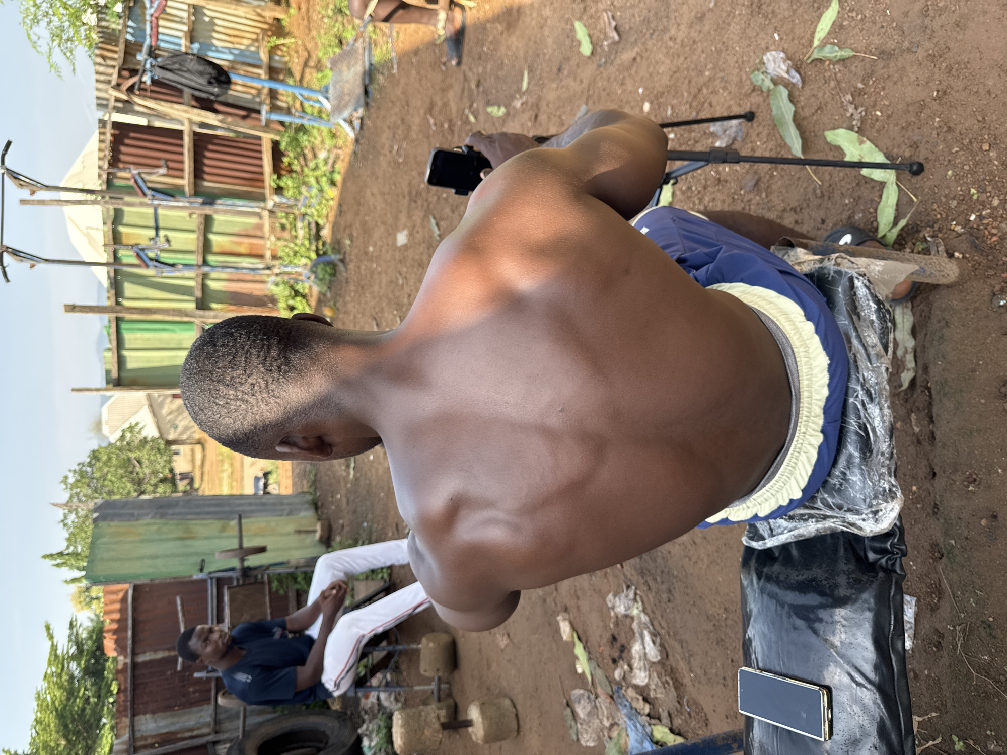 In Akwanga, Nigeria, an outdoor gym setup is depicted. In the foreground, a shirtless man with dark skin is seated facing away from the camera, his back and shoulders visible, appearing muscular. He holds a black smartphone in a black holder. To his left, another man, wearing a dark t-shirt and white pants with red stripes, is seated on a low stool, facing left. In the background, there is a collection of makeshift exercise equipment, including weightlifting bars, dumbbells, and pull-up bars, constructed from various scrap materials such as corrugated metal sheets and pipes. The ground is dirt, and scattered debris is visible, suggesting a rugged, informal environment. Sunlight illuminates the scene, casting shadows and highlighting the textures of the surroundings.