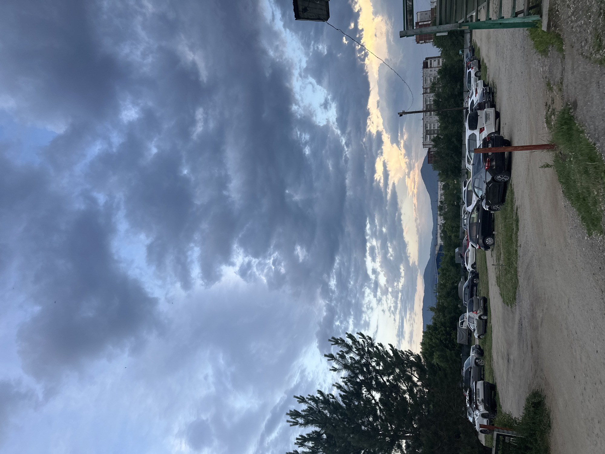 The image depicts an outdoor parking lot with several vehicles parked on a dirt and gravel surface. The parking lot is situated in front of a line of trees and buildings, with mountains visible in the background. The sky is overcast with dramatic clouds, and there is a hint of sunset light near the horizon. A large pine tree is prominent on the left side of the frame. There are no people visible in the image. The overall scene suggests a somewhat rural or suburban setting on the outskirts of a city, likely Sayanogorsk, Russia, given the typical architecture and landscape.