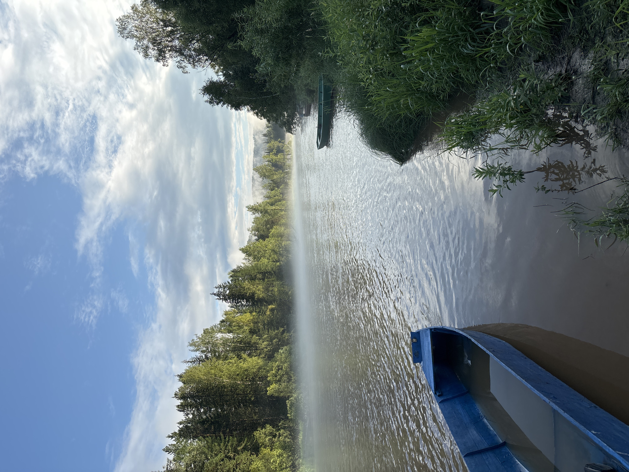 This is a medium shot of a body of water in the city of Korodovo, Russia, under a partly cloudy sky. In the foreground, on the left, is the front of a blue wooden rowboat partially submerged in the water. The boat is oriented diagonally from the lower left corner towards the center. The water has gentle ripples and reflects the sky.
The middle ground shows the river stretching into the distance, with a bank lined with dense green trees on the right. A light fog or mist hovers over the surface of the water, obscuring the far bank and distant trees. Another green rowboat is visible tied to the shore on the right. The shore itself is covered in thick green grass and vegetation.
In the background, the dense forest continues along the riverbanks, with the fog creating a hazy effect on the landscape. The sky above is a mix of blue sky and scattered white clouds. The lighting suggests it is either early morning or late afternoon. There are no people visible in the image.
