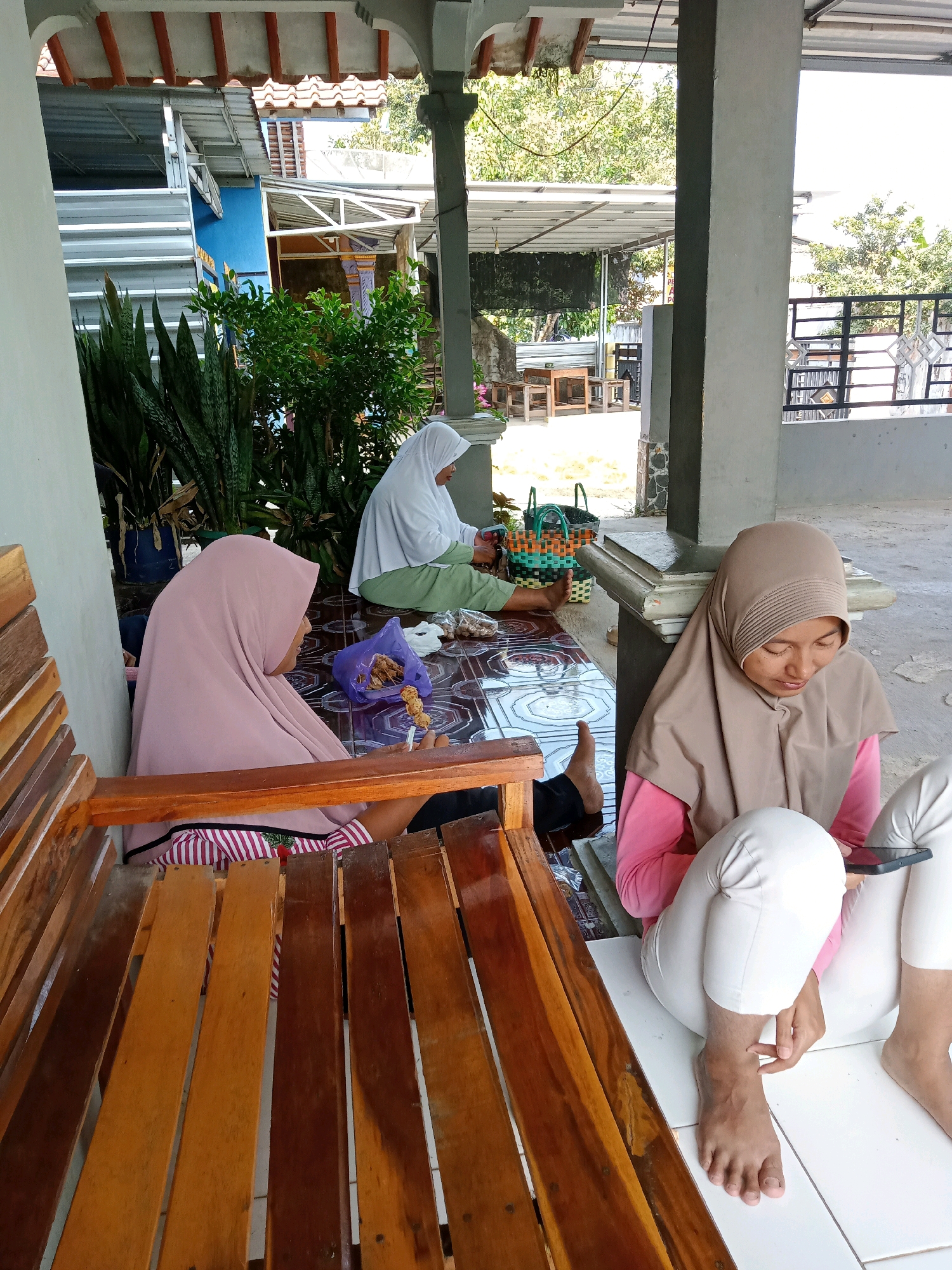 Three individuals wearing hijabs are sitting on the floor in an outdoor covered patio area, possibly in Gumiwang, Indonesia. A wooden bench is in the foreground. The woman on the left, wearing a pink hijab, is seated on the bench and holding a skewer of food. The woman in the middle, wearing a white hijab, is sitting on the floor and appears to be sorting through items in a woven basket. The woman on the right, wearing a brown hijab, is also sitting on the floor and looking at a smartphone. There are woven baskets and food items scattered on the floor. The background shows a building with a tiled roof and lush green vegetation. The lighting suggests it is daytime.