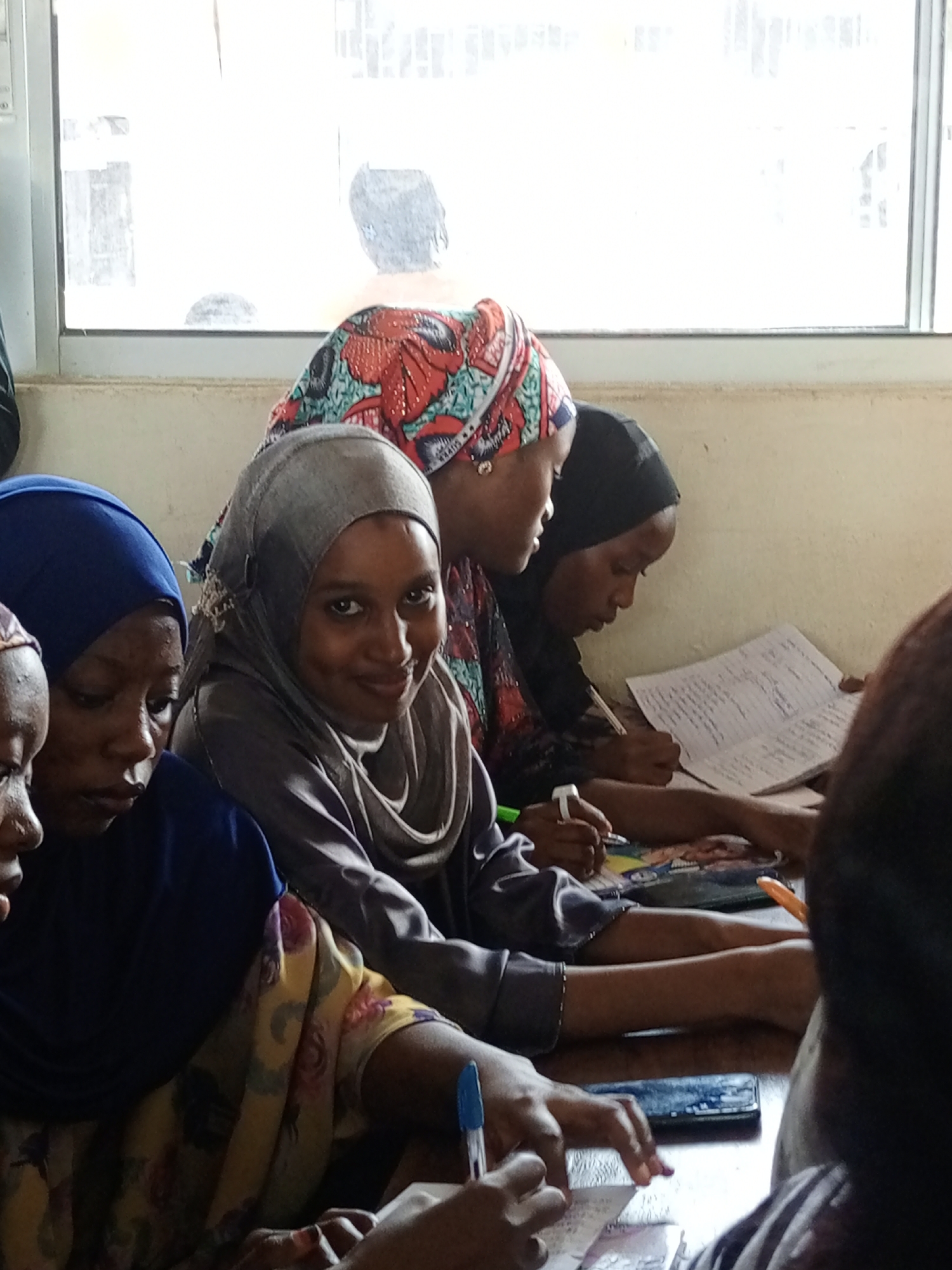 A group of women, likely young adults, are gathered indoors, possibly in a classroom or study setting in Keffi, Nigeria. The scene is brightly lit by a large window in the background, suggesting it is daytime. Most of the women are wearing hijabs or head coverings and various patterned garments.
In the foreground, on the left, a woman wearing a blue hijab is partially visible, focused downwards. Next to her, another woman in a gray hijab and gray top looks towards the camera with a gentle smile, her eyes sparkling. Her right hand is near a notebook on the table. Behind her, another woman with a colorful patterned head covering and a black top is actively writing in a notebook with a pen. Her notebook is open to a page filled with handwritten text and possibly some diagrams. To her right, another woman, mostly obscured from view, also appears to be writing.
On the table in front of them are several notebooks, pens (including a blue pen and a white-capped marker), and a dark-colored mobile phone. The women appear engaged in a learning or study activity, diligently writing. The background features light-colored walls and the large, bright window. The overall atmosphere appears focused and collaborative, with a hint of warmth from the smiling woman.