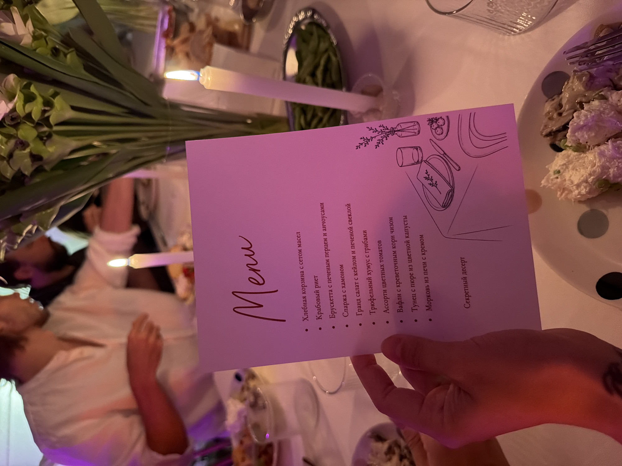 In a dimly lit indoor setting, likely a restaurant or event space, a hand holds a pink menu with "Menu" printed in elegant script at the top. The menu lists various food items in Russian, including "Хлебная корзина с сетом масел" (Bread basket with a set of oils), "Крабовый риет" (Crab rillettes), "Брускетта с печеным перцем и анчоусами" (Bruschetta with baked pepper and anchovies), "Спарка с хамоном" (Sparka with ham), "Гранд салат с кейлом и печеной свеклой" (Grand salad with kale and baked beetroot), "Трюфельный хумус с грибами" (Truffle hummus with mushrooms), "Ассорти цветных томатов" (Assortment of colored tomatoes), "Вафли с креветочным корн чизом" (Waffles with shrimp corn cheese), "Тунец с пюре из цветной капусты" (Tuna with cauliflower puree), "Морковь из печи с кремом" (Carrot from the oven with cream), and "Секретный десерт" (Secret dessert). A black and white line drawing of a table setting with cutlery and a small vase of flowers is visible on the lower right portion of the menu.
The background shows blurred figures of people dining, with a large bouquet of green flowers and white calla lilies in a vase placed on the table. Two white candles in glass holders are lit, casting a warm glow. In the foreground, a plate of food, appearing to be a salad or appetizer with creamy components and mushrooms, is partially visible, along with a fork. The overall ambiance suggests a formal or celebratory meal. The image does not provide any specific geographical indicators to place it in Chang Phueak, Thailand.