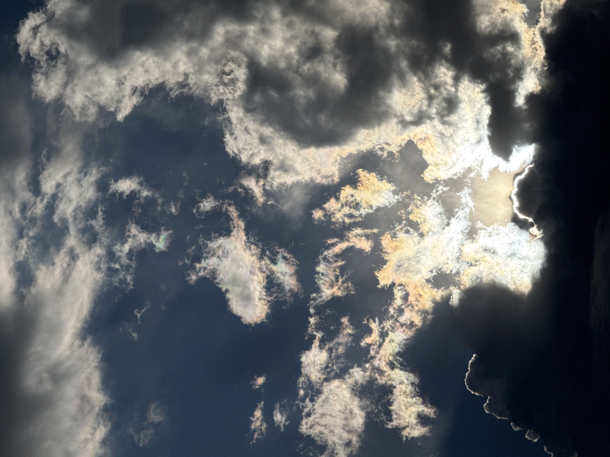 This is a photograph of a cloudy sky, taken from below, looking upwards. The clouds are varied in density and color, with some appearing dark and ominous, while others are brightly lit by the sun, creating a dramatic contrast. The sun is partially obscured by a large, dark cloud formation on the right side of the frame, but its intense brightness can be seen peeking through, casting rays of light that illuminate the edges of other clouds with a golden and iridescent glow.
The clouds exhibit a range of textures, from wispy and thin to thick and voluminous. Some clouds have sharp, defined edges, particularly where the sunlight catches them, while others appear softer and more diffused. The sky itself is a deep blue, suggesting it is daytime. There are no people or man-made objects visible in the image, focusing solely on the natural phenomenon of the sky and its atmospheric conditions.
The lighting suggests it might be late afternoon or early evening, as the sun's angle and the intensity of the light create strong shadows and highlights. The overall mood is dramatic and awe-inspiring, with the powerful interplay of light and shadow in the clouds evoking a sense of grandeur and the raw power of nature. The location context of Pavlodar, Kazakhstan, does not provide any specific visual cues within the image itself, as the scene is a universal depiction of a cloudy sky. There is no visible text in the image.