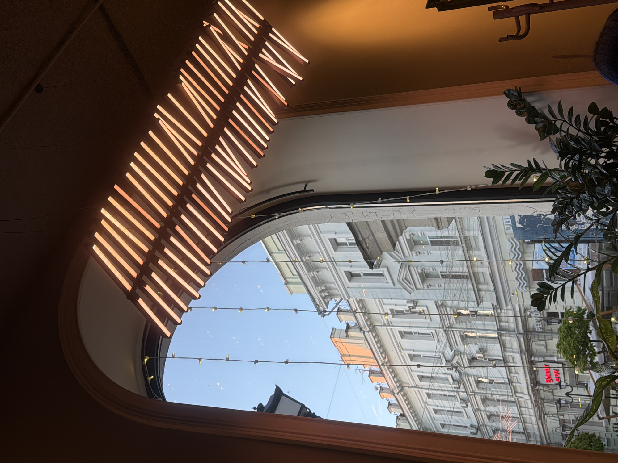 The image captures an interior view looking out through a large, arch-shaped window onto a city street. Dominating the upper left is a modern light fixture, an artistic arrangement of numerous parallel glowing strips, likely neon or LED, emitting a warm orange-red light. The light fixture is angled and appears to hang from a dark ceiling.
Through the window, a bright blue sky is visible, along with reflections of the interior ceiling and the light fixture itself. The exterior view reveals multi-story buildings with detailed architecture, numerous windows, and decorative elements, suggesting an urban setting. Strings of small, glowing fairy lights are strung across the window opening, adding a decorative touch. On the right side, a dark green leafy plant, possibly an indoor plant, is partially visible, framing the outdoor view.
The scene appears to be set during daytime, under clear and sunny weather conditions. The architectural style of the buildings outside suggests an older urban environment, possibly in a European city. A notable detail in the reflection on the window glass is inverted red text from a sign, which clearly spells "РЕСТОРАН" (RESTORAN), meaning "restaurant" in Russian. This indicates that the interior space might be a restaurant or café. The overall atmosphere is bright and inviting, blending contemporary interior design with a classic city outlook.