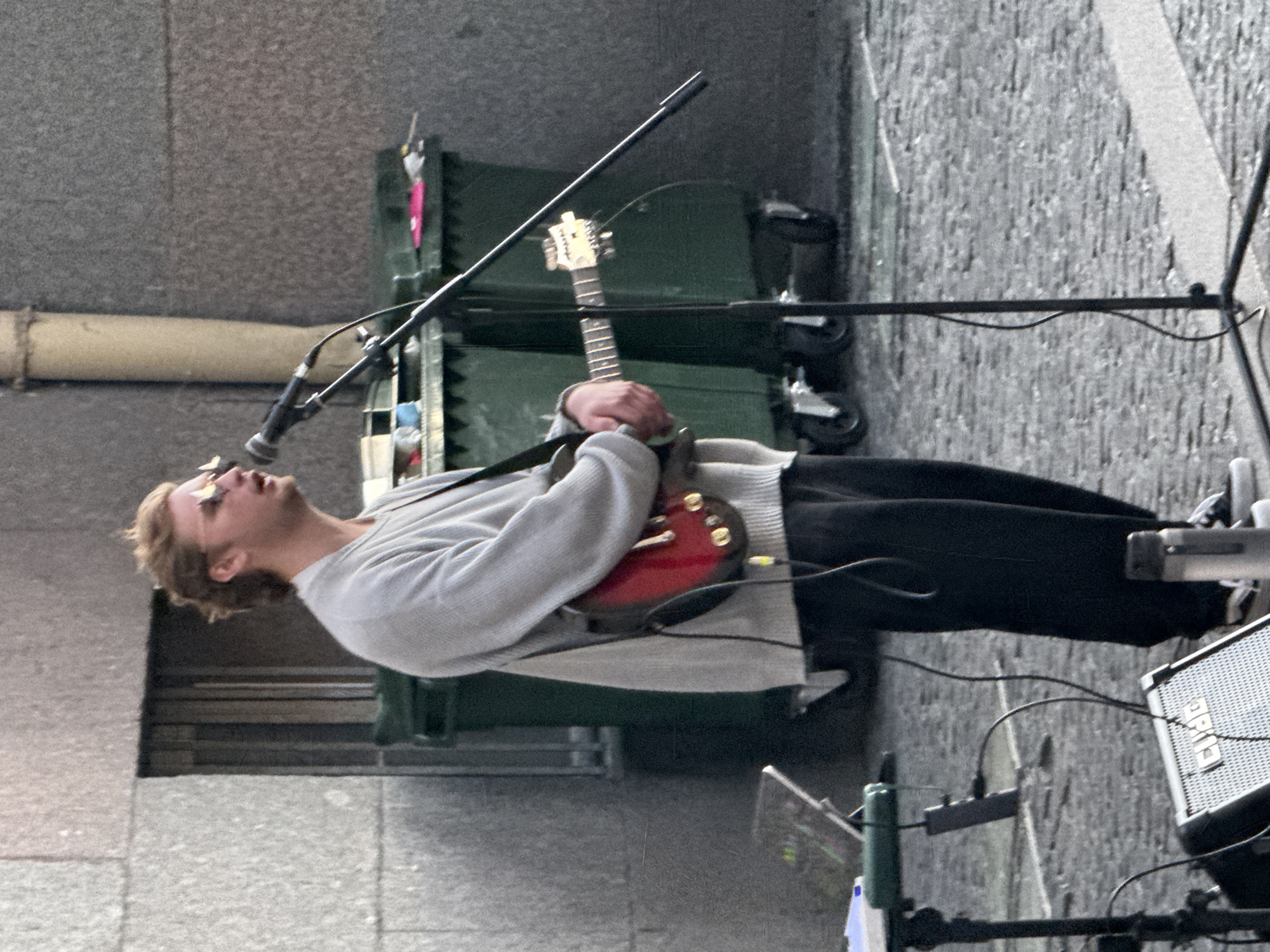 A light-skinned male with blonde hair, wearing a light gray sweater, dark pants, and sunglasses, stands outdoors in what appears to be Saint Petersburg, Russia. He is holding a red electric guitar and singing into a microphone. The background shows a stone building facade, two large green dumpsters, and a cobblestone street. Equipment, including a music stand and a small amplifier with the logo "CUBE" visible, is set up around him, suggesting he is performing as a street musician. The lighting indicates it is daytime, possibly overcast.