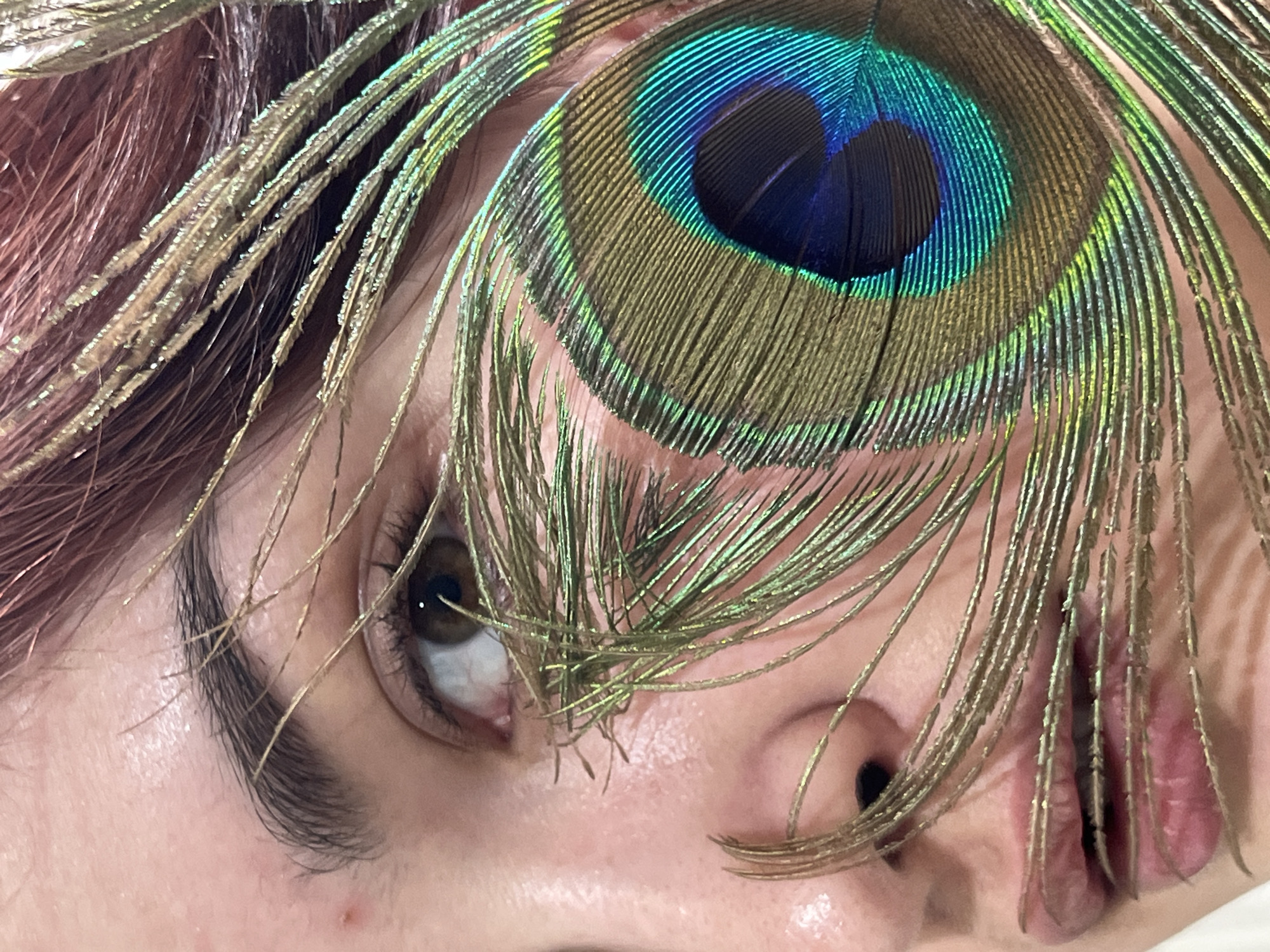 A close-up, vertical photographic portrait depicts the right side of an individual's face, located in Neftekamsk, Russia. The subject has fair skin, with a minor blemish observed superior to the right eyebrow. The right eye is open, displaying a dark brown iris and a visible pupil, directed slightly supero-laterally. Dark, defined eyelashes are present. The lips are slightly parted. Reddish-brown hair is visible along the superior-right frame edge. A peacock feather is positioned over the face. Its prominent iridescent ocellus, featuring concentric rings of blue, green, and gold, is located inferior and lateral to the right eye, partially covering the cheek. The feather's filamentous barbs extend across the right eye, the nasal bridge, and obscure portions of the upper and lower lips. The immediate background is a uniformly light, out-of-focus surface.