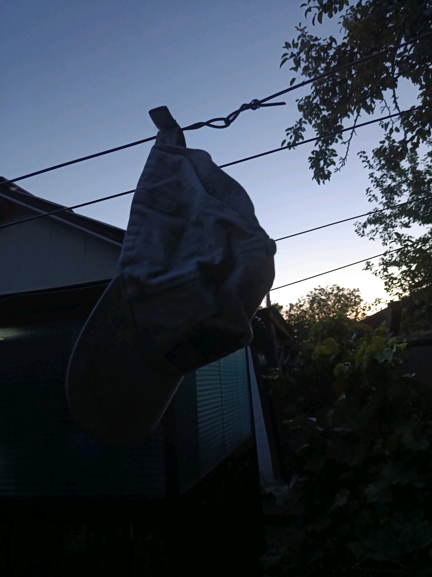A grey baseball cap is hanging on a wire, suspended by a knot in the wire. The cap is silhouetted against a pale blue sky, with the sun beginning to set behind trees in the background. The cap is partially obscured by a dark green leafed tree on the right. The cap is also hanging above a dark green building, which appears to be a residential building in Smila, Ukraine. The overall mood of the image is peaceful and serene.