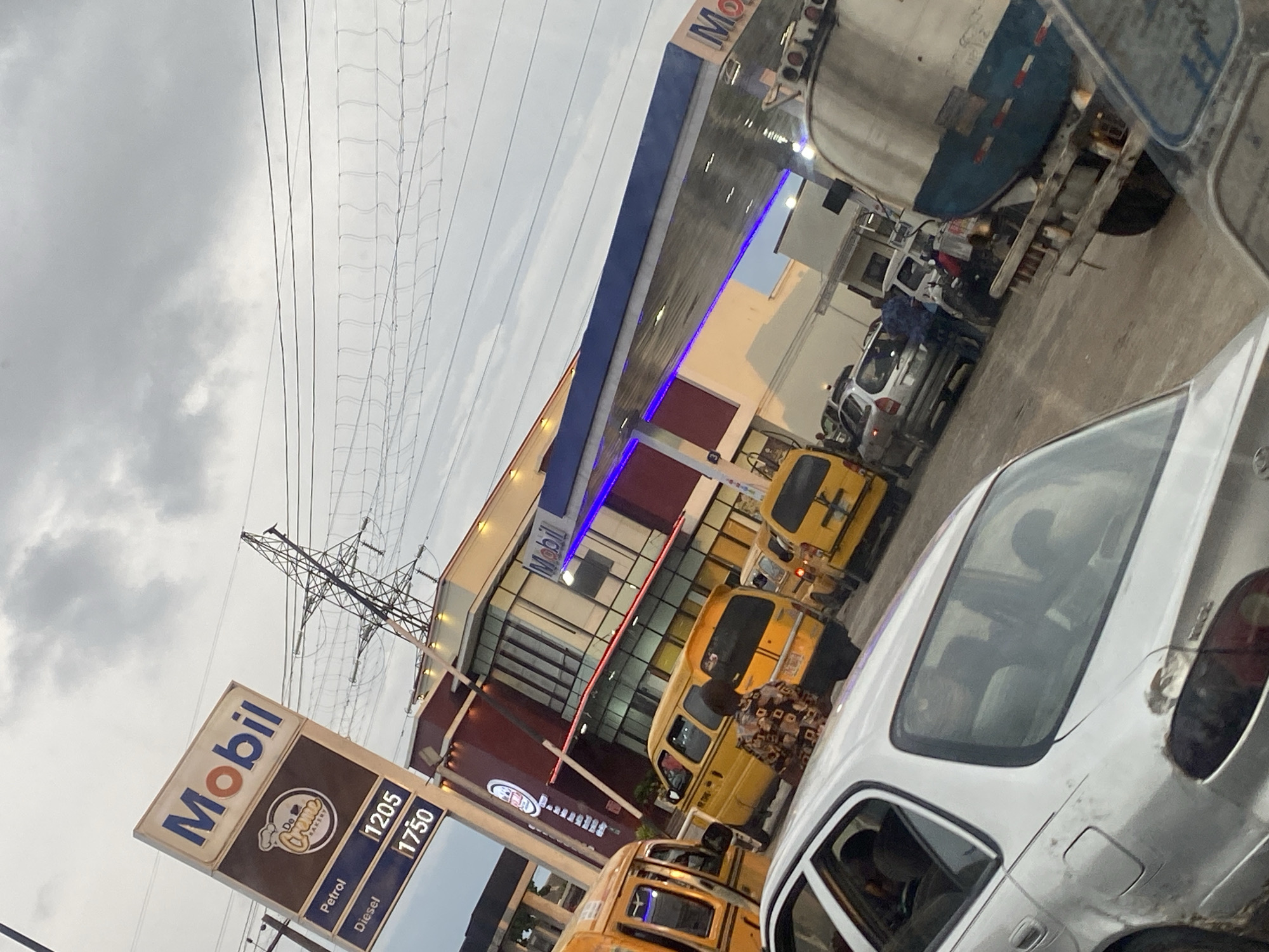 This is a medium shot, taken from a slightly low angle, of a busy Mobil petrol station in Lagos, Nigeria. The scene is dominated by a long queue of vehicles, including yellow minibuses and cars, waiting to refuel. A white car is in the foreground, and to its right, a large tanker truck is parked. Behind the vehicles, the modern Mobil station building with its distinctive blue canopy and red accents is visible. A tall electrical pylon looms in the background against a cloudy sky. The atmosphere appears to be one of typical urban congestion, with the gas station serving as a focal point for vehicular activity.