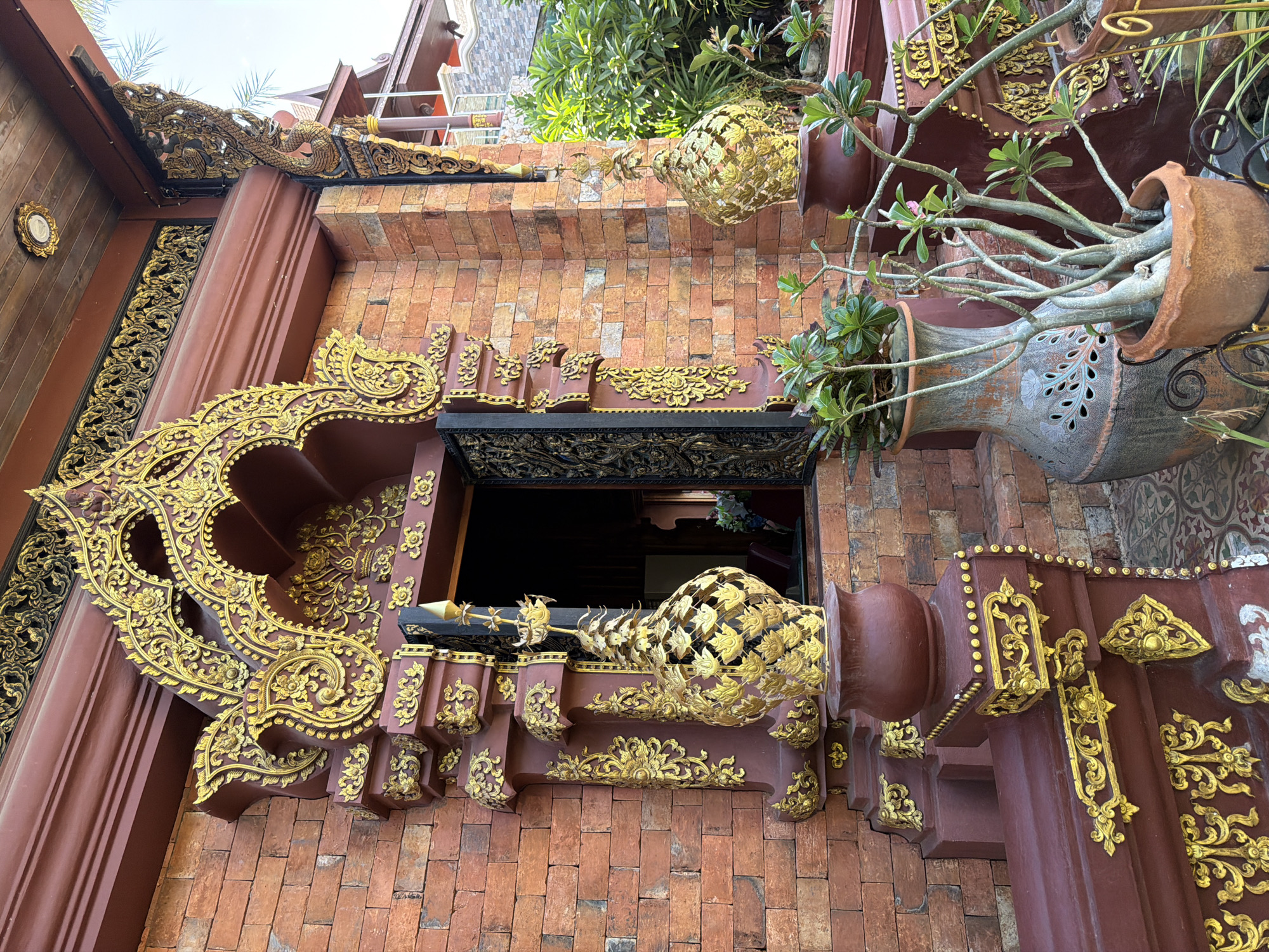The image shows the ornate entrance to a building constructed of reddish-brown brick. The entrance is framed by a dark red structure adorned with intricate gold detailing in floral and scrolling patterns. Above the entrance, a large, elaborately carved gold archway is visible. To the left and right of the entrance are large, dark red pedestals topped with gold decorative elements that resemble stylized urns with lotus flower motifs. In front of these pedestals, on the ground, are several potted plants. The plant in the foreground is in a terracotta pot and has thick, succulent-like stems and sparse green leaves, with a few small pink flowers. Behind it is a large, grey ceramic vase with cut-out floral designs. A wooden ceiling is visible above the entrance, with a decorative gold fixture attached to it. To the right of the entrance, a glimpse of palm trees and other greenery can be seen, suggesting an outdoor setting with a bright sky. The overall impression is one of cultural or religious architecture, likely in a warm climate.