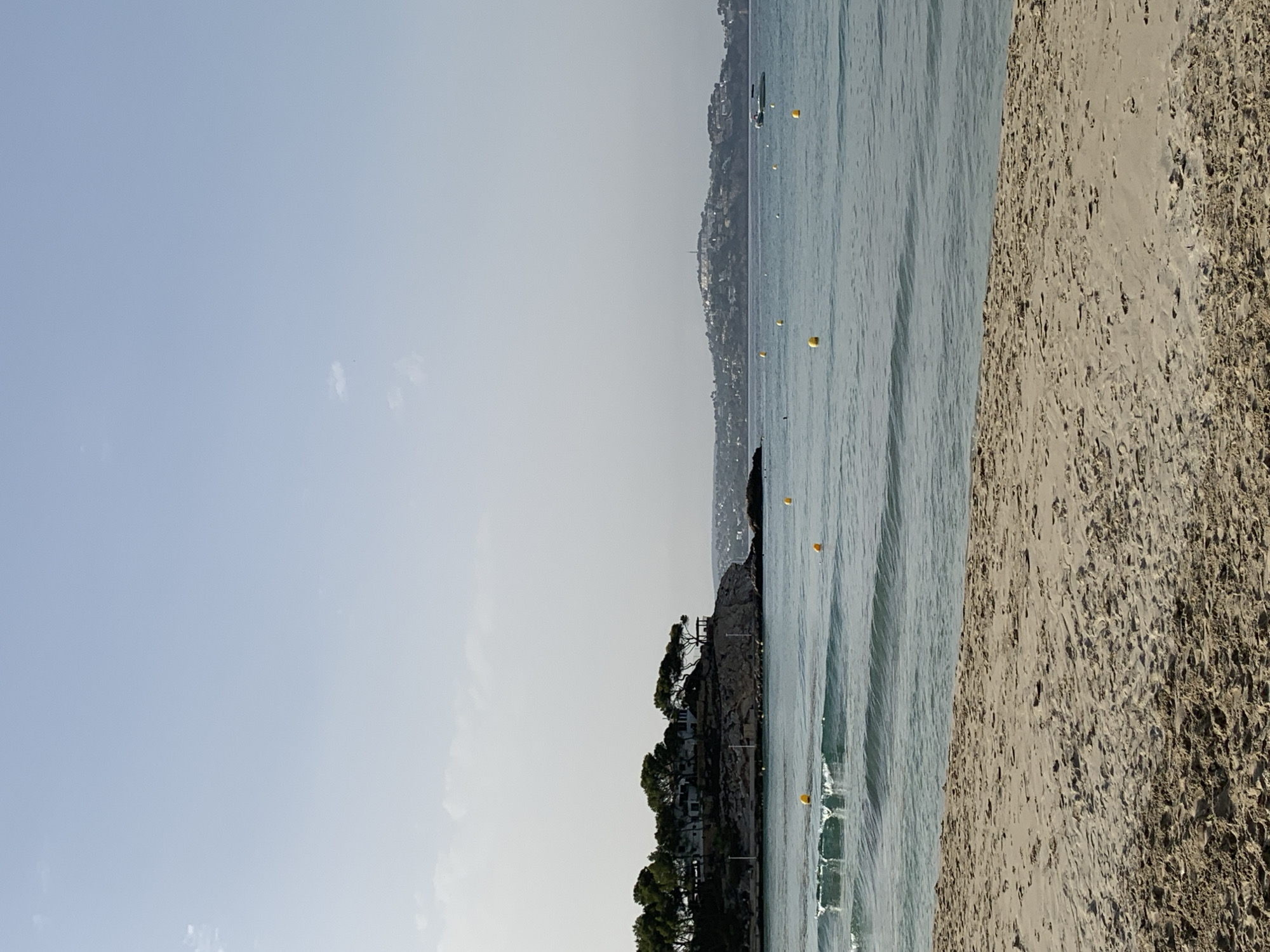 The image shows a serene beach scene in Calvià, Spain, likely in the morning or late afternoon given the soft lighting. The foreground is dominated by a sandy beach with subtle ripples and footprints indicating recent activity. Gentle waves with white crests roll towards the shore, creating a calming rhythm. Scattered across the water are several bright yellow buoys, marking a designated area or warning.
In the mid-ground, a rocky outcrop juts into the sea, topped with a cluster of lush green pine trees and some white structures, possibly homes or a resort. The architecture suggests a Mediterranean style, blending with the natural landscape. A small gazebo or shelter is visible on the edge of the outcrop.
Further in the distance, across the tranquil blue water, a hilly coastline rises, dotted with buildings, forming a settlement that extends up the slopes. A small white boat navigates the calm sea towards the right side of the frame. The sky is a pale, clear blue with a few wispy clouds, contributing to the overall peaceful atmosphere. The scene captures a typical coastal view in this popular tourist region of Mallorca.
