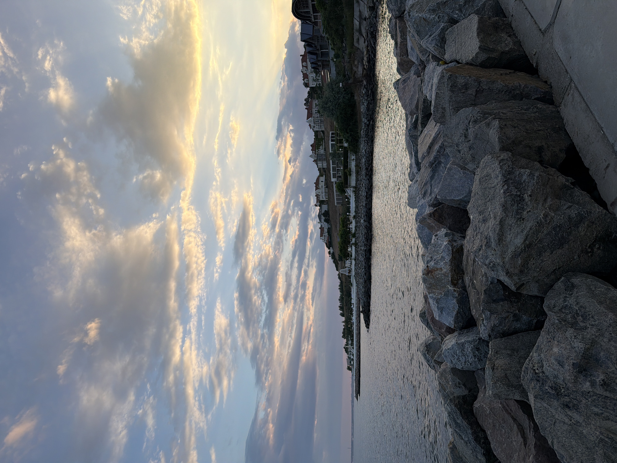 The image captures a twilight view of the coast in Fontanka, Ukraine. In the foreground, large, irregularly shaped grey rocks form a seawall, with a paved walkway visible to the right. Beyond the rocks, a body of calm water stretches towards the horizon, reflecting the muted colors of the sky. The middle ground features a picturesque coastline dotted with houses and trees situated on a gentle slope. The sky above is a canvas of soft, diffused light, with wispy clouds tinged with hues of pink, orange, and blue, suggesting the lingering presence of the setting or rising sun. No people or animals are visible in the scene.