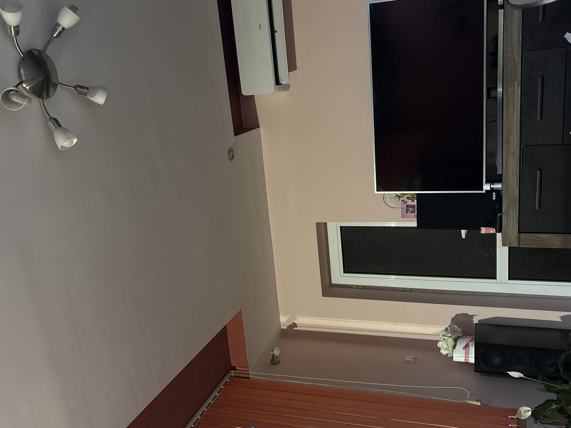 The image displays the interior of a room, likely a living area, characterized by a dual-tone wall color scheme featuring light peach/pink and light gray/white sections.
A modern, brushed silver ceiling light fixture with five small white, conical shades is mounted on the light gray ceiling, though one bulb is exposed from a missing shade. High on the peach-colored wall, a white wall-mounted air conditioner unit with "Fujitsu" branding is visible. Adjacent to the air conditioner, a large flat-screen television with a black screen and silver bezel is positioned vertically (when corrected for orientation), next to a dark wooden cabinet with dark drawers. Below the television, a black audio component bearing "B&W" text is present.
Further down the wall, a window fitted with dark roller blinds or shades is framed by white casing. Directly below the window, a black speaker or soundbar sits on a surface. To its left, a small white gift box adorned with a pink ribbon and artificial flowers is placed. A green plant with a white flower, resembling a Peace Lily, is situated to the right of the speaker. The lower section of the wall features horizontal wooden blinds, partially obscured by a transparent barrier, possibly acrylic, supported by small metallic fixtures.
The room is illuminated by artificial light, suggesting either evening or night, or that exterior light is blocked by the blinds. There are no people visible, and no specific activities are taking place.