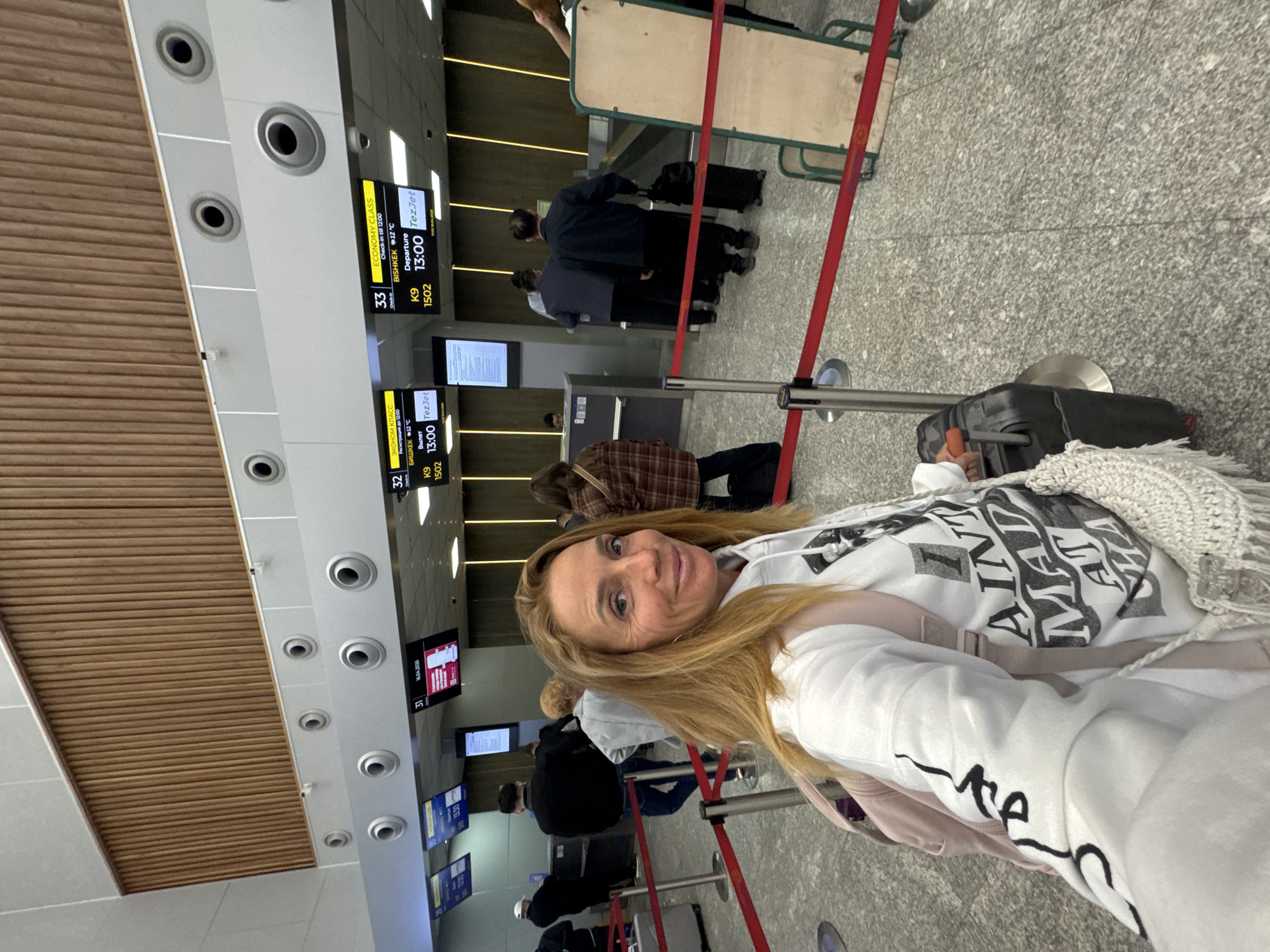The image shows a woman in an airport. The woman is blonde and is wearing a white sweatshirt. She is standing in a line, likely for check-in. In the background, there are monitors displaying flight information, including details for flight K9 1502 to Bishkek departing at 13:00 on TezJet. There are other people in line with luggage. The environment appears to be a modern airport terminal, with a wooden ceiling and gray flooring. There is no indication that the image was taken in the city of Tashkent, Uzbekistan.