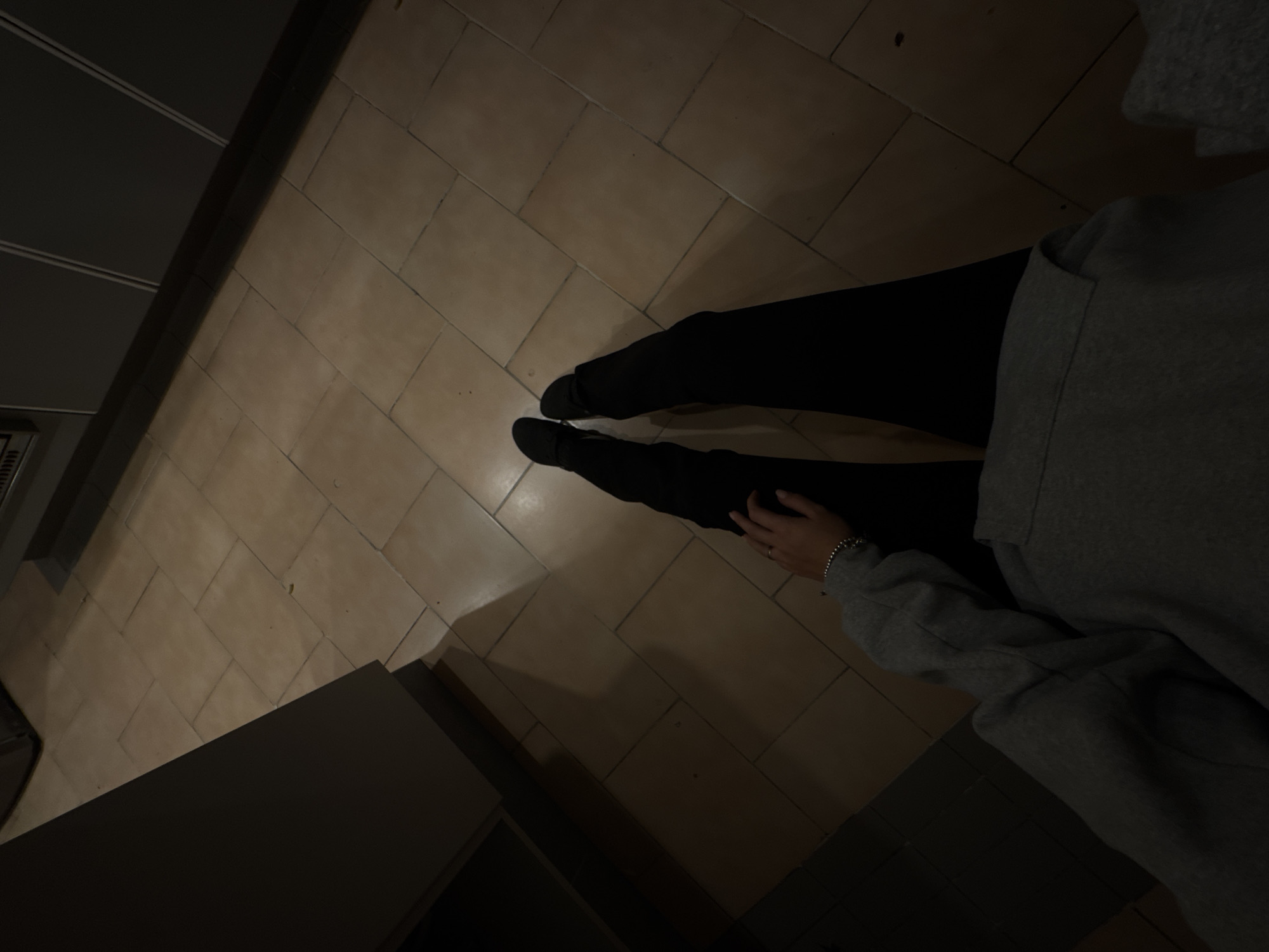 The image depicts a person sitting on a tiled floor, wearing black pants and a grey hoodie. The setting appears to be indoors, potentially a kitchen or similar space with built-in cabinetry visible on the left side of the frame. The tile flooring is a light brown color with rectangular patterns. The lighting is dim, creating strong shadows throughout the scene. A hand is resting on the person's leg, with a silver bracelet visible on their wrist. The exact location in Mexico City, Mexico, is indeterminable based on the provided image.
