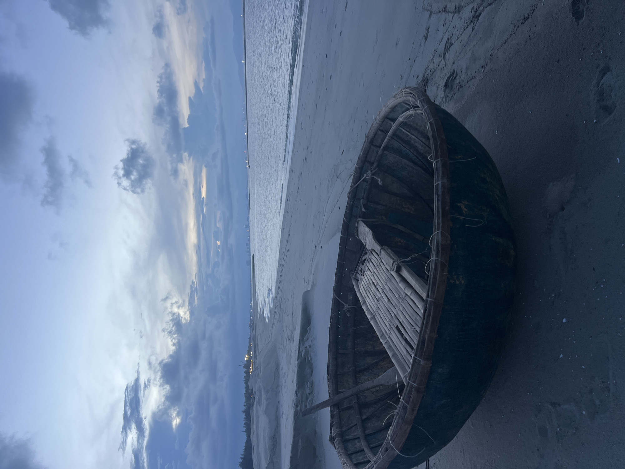 A wide-angle, eye-level shot captures a round, dark blue coracle boat resting on a sandy beach in Hoi An, Vietnam, during twilight. The boat, weathered and rustic, features a simple bamboo seating arrangement inside and a wooden oar. The calm, rippling sea stretches out to the horizon, reflecting the overcast sky filled with dramatic, dark clouds. In the distance, the faint lights of a city or port town are visible along the coastline, with a long, thin structure extending into the water. The sand is a light grey, showing subtle footprints and tire tracks. The overall mood is serene and slightly melancholic, indicative of the transition from day to night.