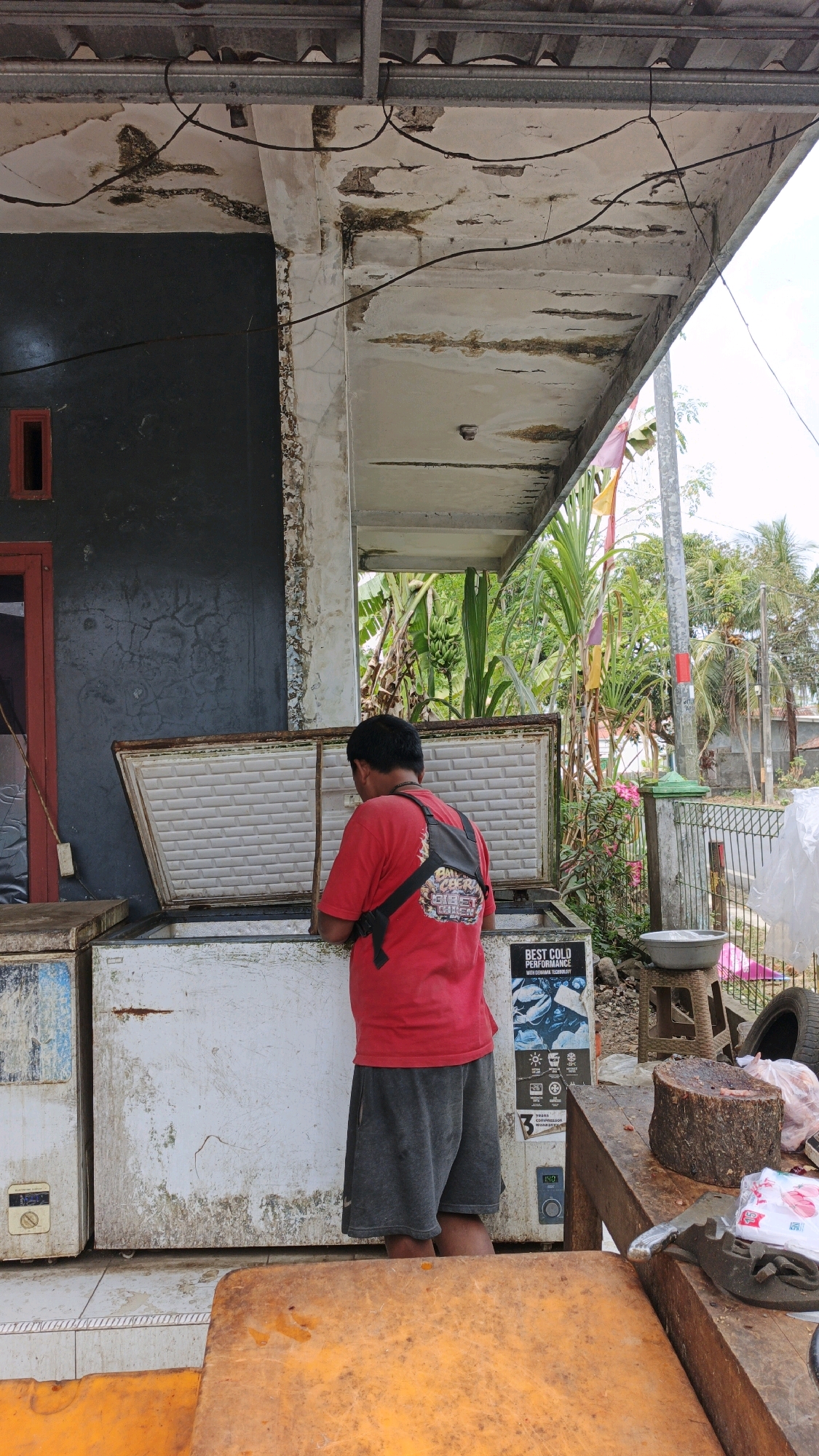 A man wearing a red t-shirt and grey shorts is standing in front of an open chest freezer. The freezer is white and appears to be old and dirty, with some rust around the edges. The man has a black backpack on his shoulders. He is reaching into the freezer with his right hand, holding a wooden stick. In the background, there is a building with a dark grey wall and a red-framed window. Above the building, there is a corrugated metal roof with some peeling paint and exposed wiring. Beyond the building, there are green plants, a street light, and a fence. On a wooden table to the right of the freezer, there is a large piece of meat on a chopping block, some plastic bags of meat, and a small metal tool. A sticker on the freezer reads "BEST COLD PERFORMANCE." The scene appears to be an outdoor market or a shop in Cibenda, Indonesia, given the presence of tropical plants and the architectural style.