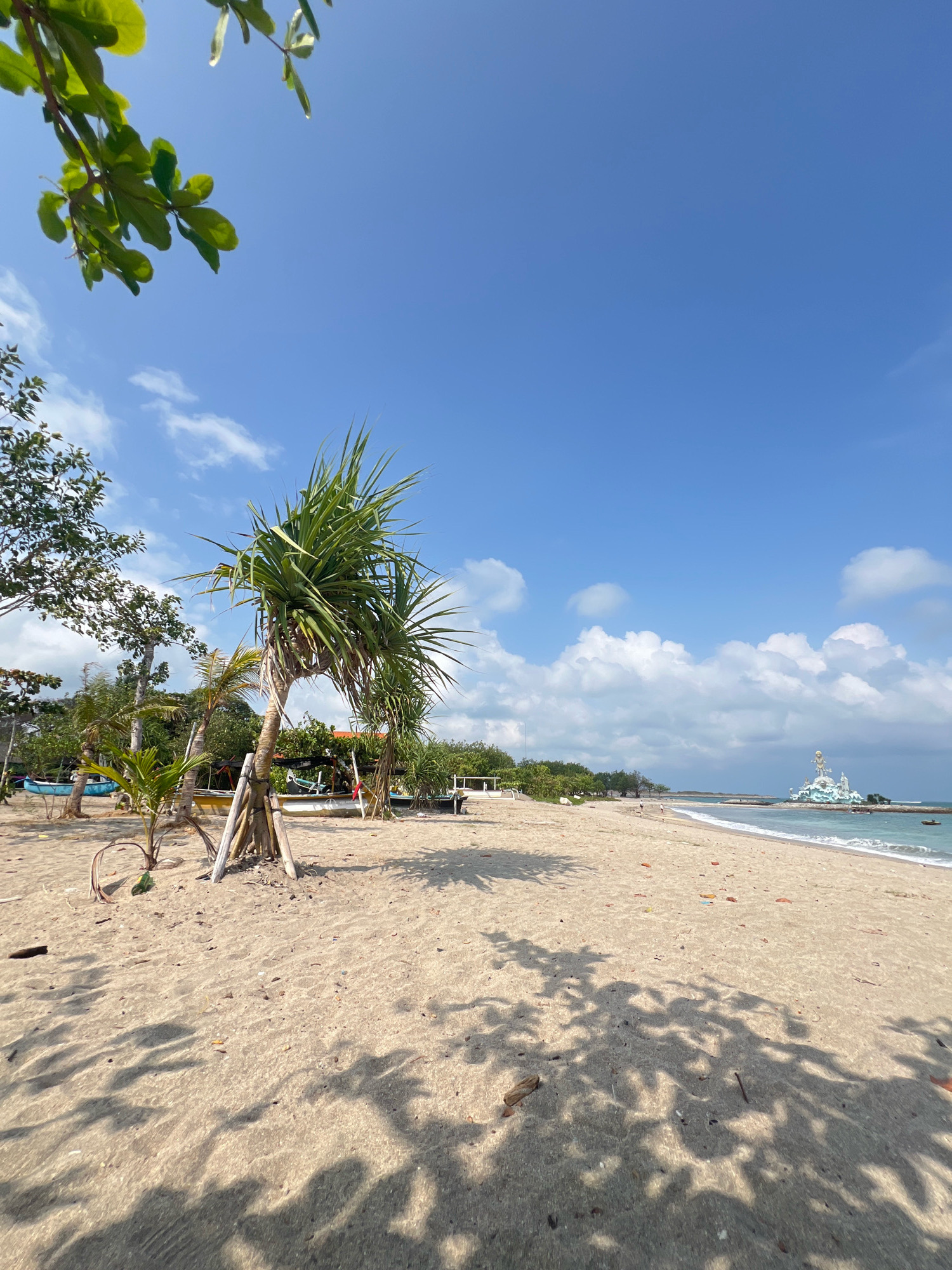 A serene beach scene unfolds under a clear blue sky with scattered white clouds. The foreground is dominated by the warm, sandy expanse of Kuta Beach, Indonesia, with scattered debris and the dappled shadows of foliage suggesting the presence of trees nearby. In the mid-ground, several small boats are pulled up onto the shore, and a distinctive pandanus plant stands prominently, supported by bamboo poles. Further down the beach, a white, multi-tiered structure, possibly a monument or building, is visible in the distance, situated on a rocky outcrop at the water's edge. The gentle waves of the ocean lap the shore, creating a tranquil atmosphere.