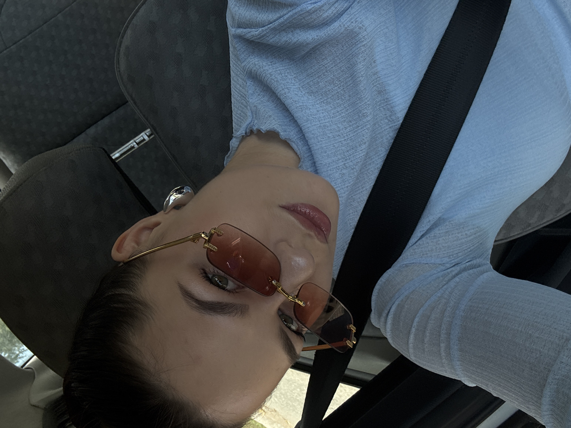 A young woman with her hair pulled back is in the passenger seat of a vehicle, wearing a light blue, textured, long-sleeved shirt, gold-rimmed rectangular sunglasses with brown lenses, and a large silver hoop earring. She is wearing a black seatbelt, which is visible across her chest. The interior of the car is dark gray with textured upholstery. The background is blurred but appears to be an outdoor scene with some foliage, suggesting the car may be parked or moving in an area with trees. The image was taken from a high angle, looking down at the woman. No specific location in Chaikovsky, Russia, can be identified from the image.