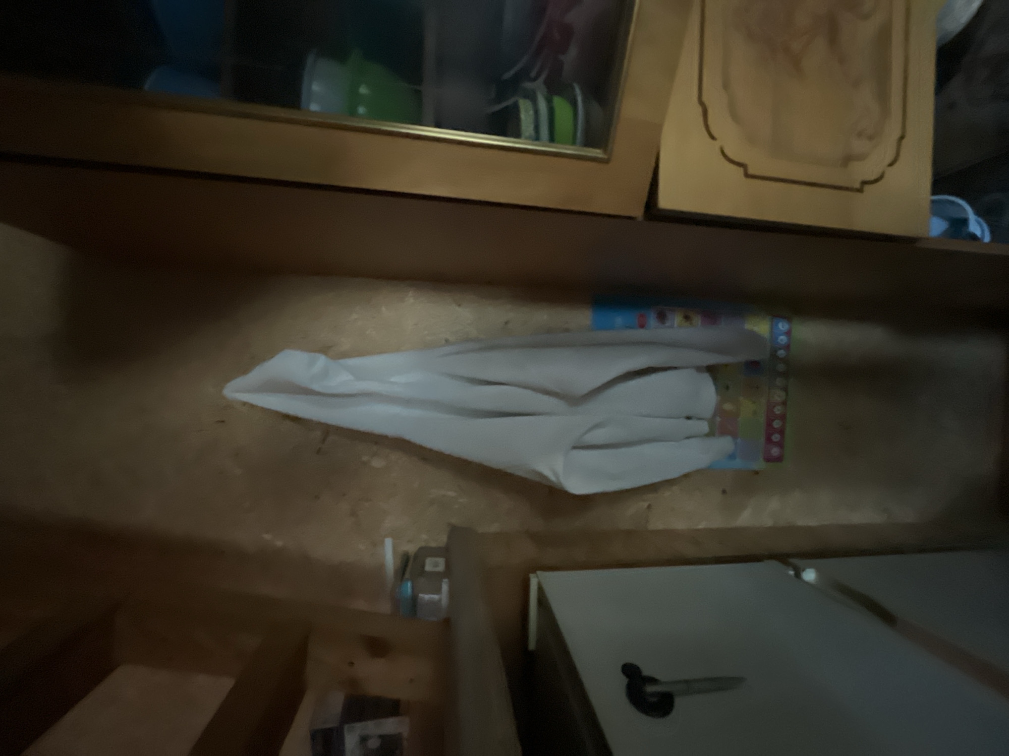 This is a medium shot, taken indoors, of what appears to be a pantry or storage area. The walls are paneled with unfinished wood, and there are several objects visible, including a white garment hanging on the wall, a shelf with small items, a cabinet, and a glass-fronted cupboard filled with containers. The lighting is dim and appears to be coming from an unseen source, casting shadows throughout the room. The overall impression is one of a functional, somewhat cluttered space.
Based on the provided crops, I cannot determine the location of this image in Chaikovsky, Russia.
