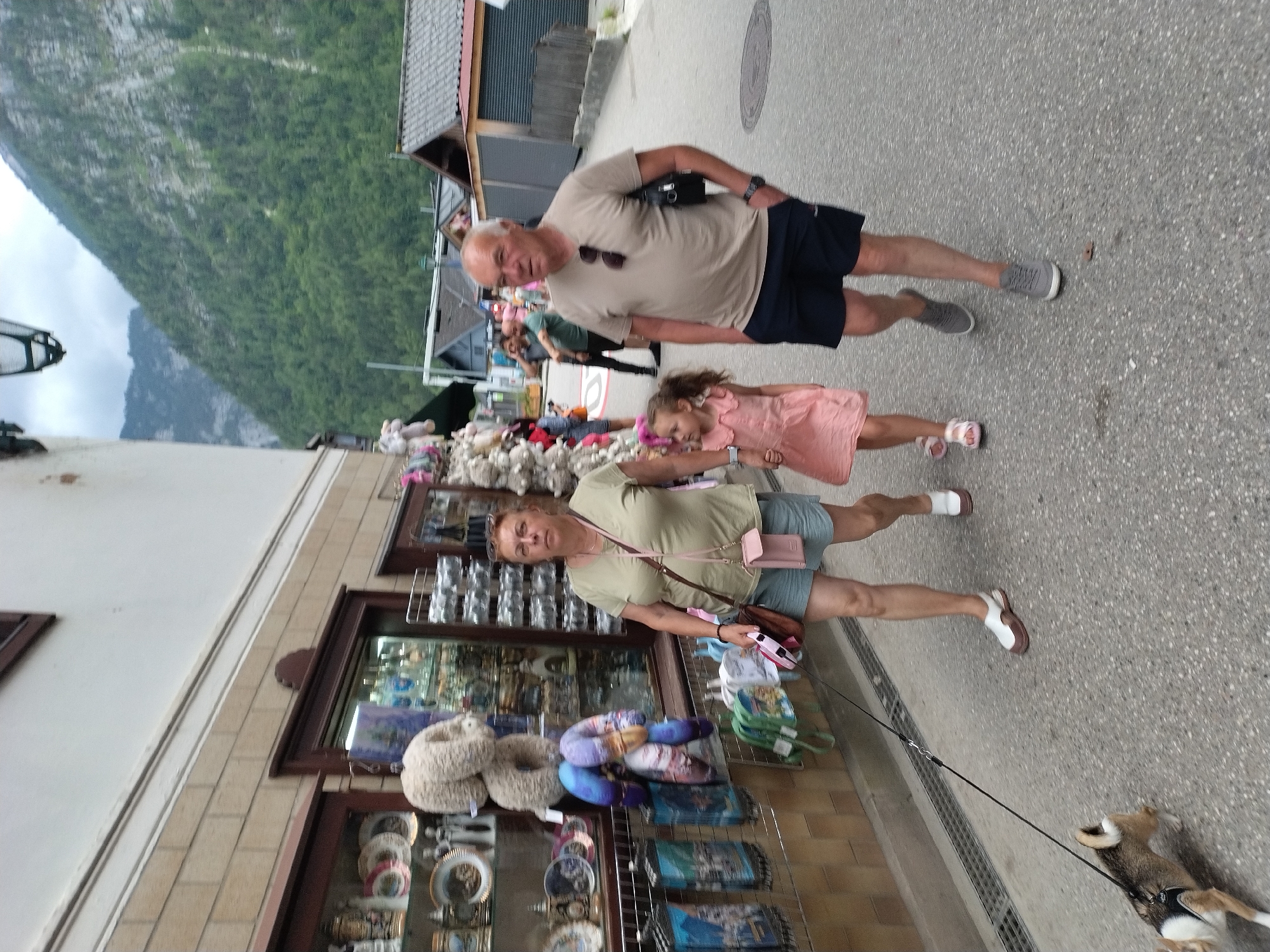 In Hallstatt, Austria, on a cloudy day, a family consisting of a man, a woman, and a young girl are walking down a cobblestone street. The woman is on the left, wearing a light olive t-shirt and blue shorts, with a brown purse slung across her body and a small pink wallet attached to her belt. She is holding a dog on a leash, and the dog is in the lower left corner of the image, walking ahead of her. The young girl, in a pink dress, walks to the right of the woman, holding her hand. The man is on the right, wearing a beige t-shirt, dark blue shorts, and grey sneakers. He has sunglasses on his shirt collar and a black bag slung over his shoulder. In the background, there is a building with shops displaying various items, including souvenirs and plush toys. The street is lined with these shops, and behind them, the steep, tree-covered mountains characteristic of Hallstatt rise up, with a glimpse of a cloudy sky at the top. Other people can be seen further down the street in the background.