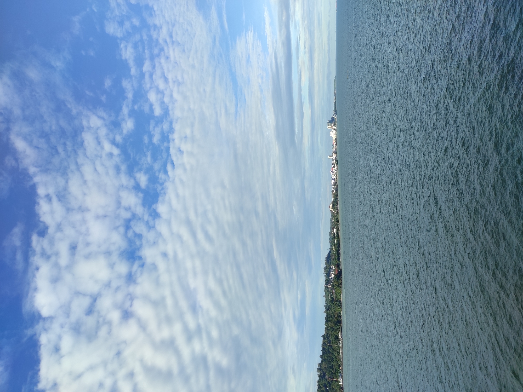 The image shows a panoramic view of the sea and the coastline of Penajam, Indonesia. The foreground is dominated by the calm, dark blue waters of the sea, with subtle ripples indicating a gentle movement. In the midground, a lush, green coastline stretches across the horizon, dotted with trees and vegetation. Along the coastline, a cityscape is visible, characterized by several buildings, including some taller structures that appear to be part of an industrial or commercial area. The sky above is a vibrant blue, filled with scattered white clouds that form interesting patterns. The overall impression is one of a serene coastal landscape with a developed urban area bordering the sea.