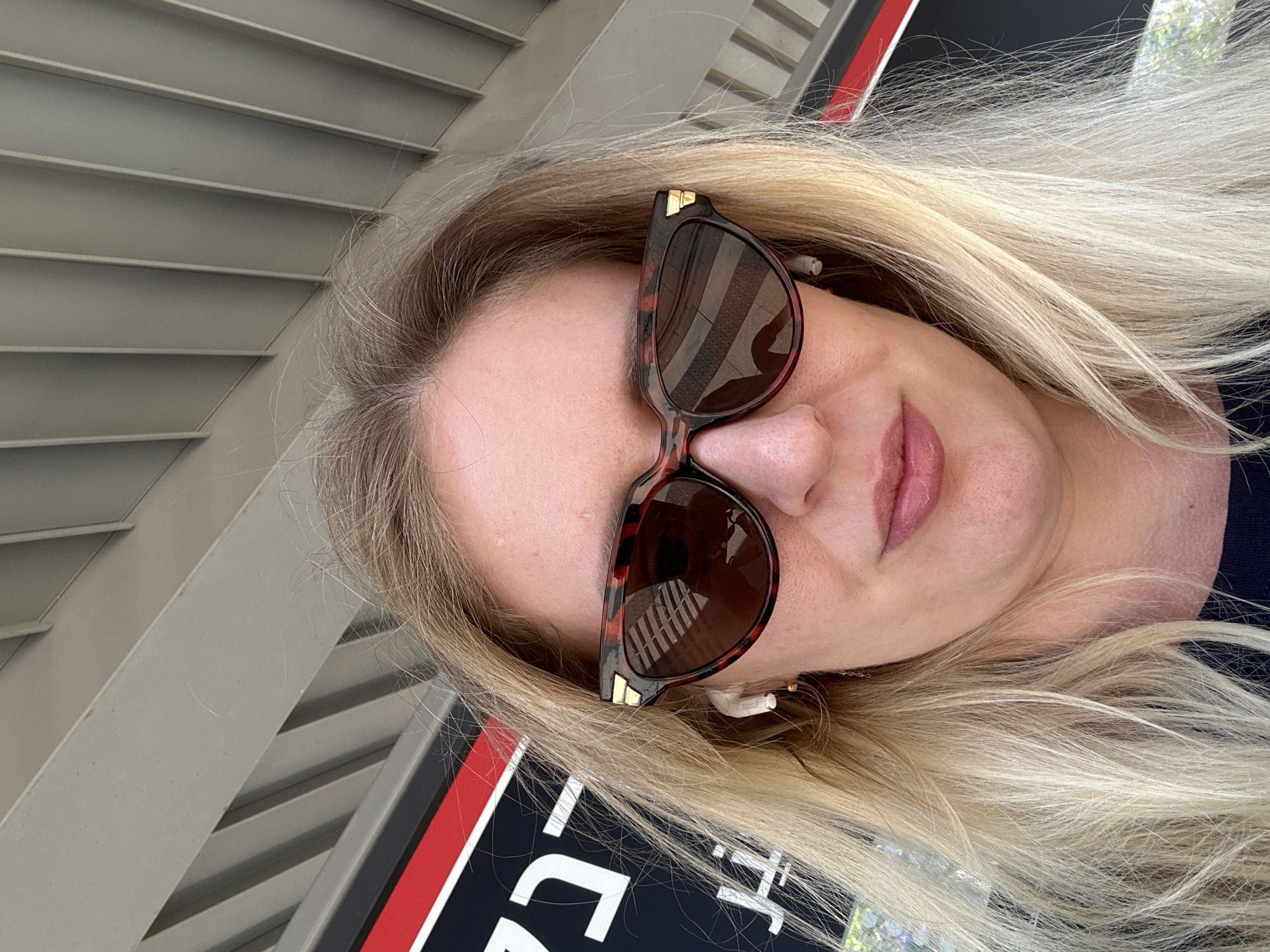 A selfie photograph shows a woman with blonde hair and sunglasses. The sunglasses have a tortoiseshell pattern. The woman is wearing wireless earbuds in her right ear. The background includes a building with horizontal blinds and a sign with black, red, and white colors. The photo doesn't depict information to find the location in the city of Petah Tikva, Israel.