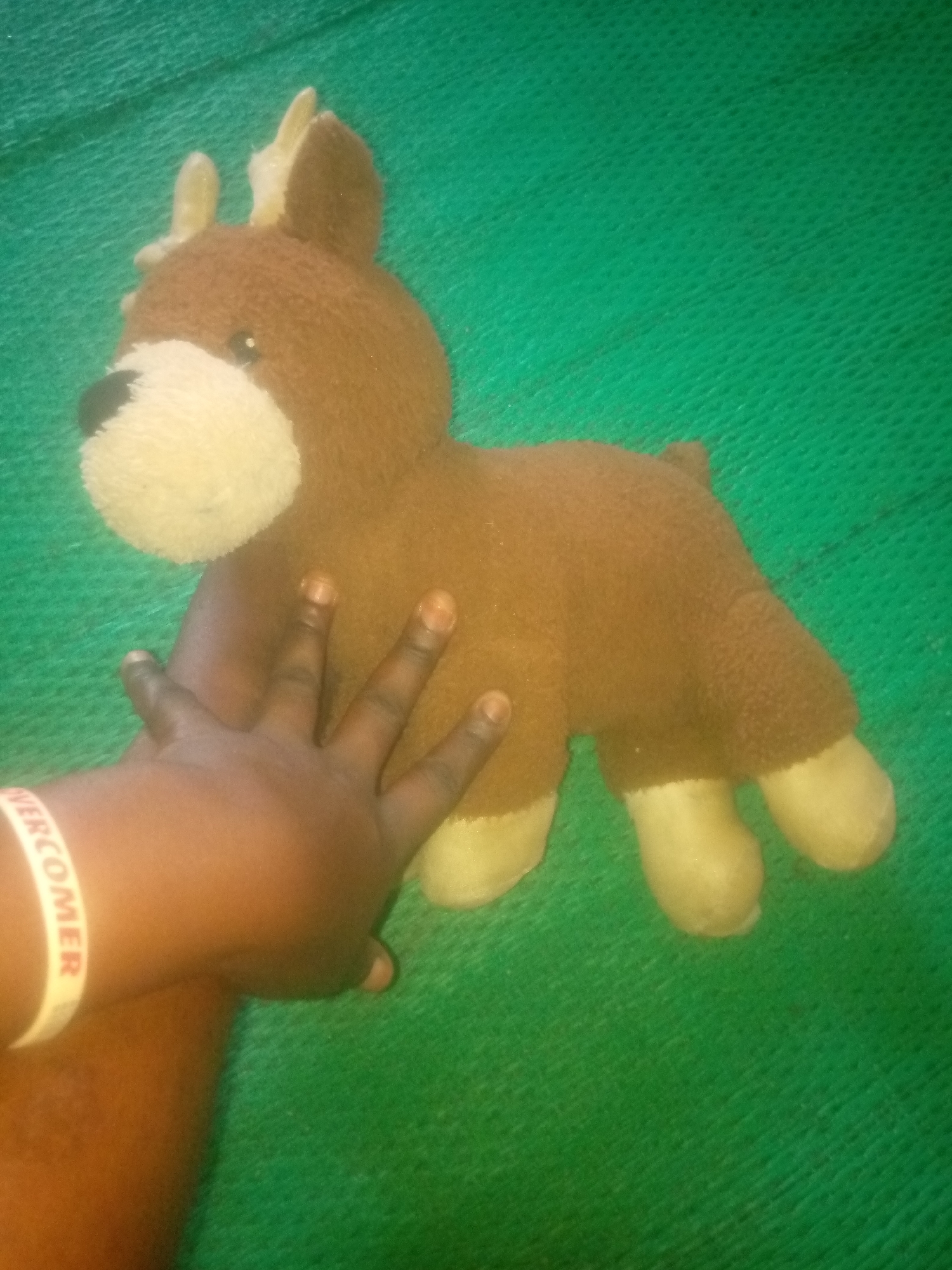A stuffed toy reindeer is held by a person's hand. The toy is brown with a white muzzle and yellow antlers. The background is a textured green surface. A white wristband with the word "OVERCOMER" is visible on the person's wrist. The scene is indoors, in an unknown location in the city of Unknown, Unknown.