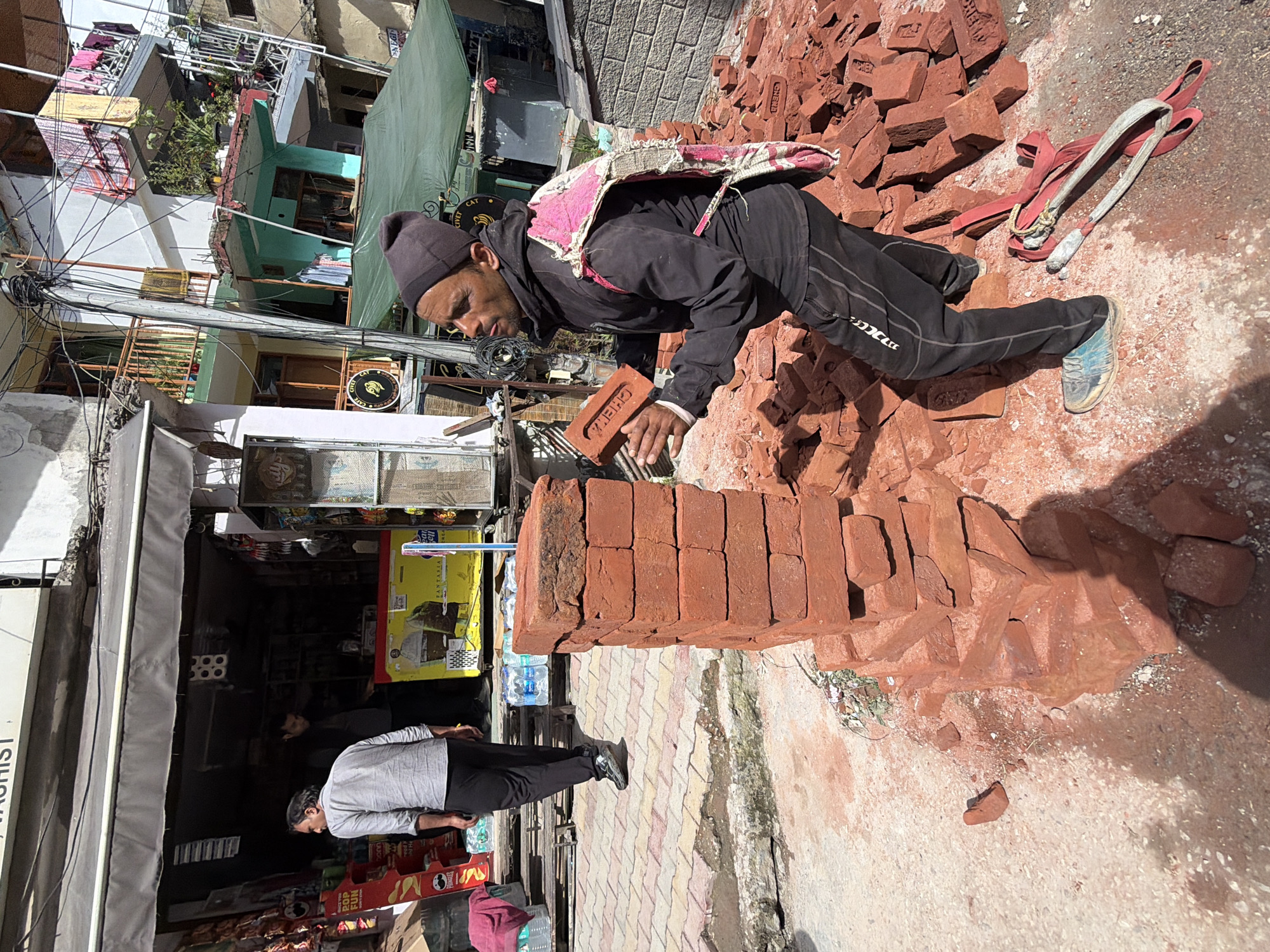 In Vashisht, India, a man wearing a dark beanie, black jacket, and black sweatpants with white stripes is in the process of stacking red bricks. He is holding one brick in his left hand and appears to be placing it onto a growing pile. He is also carrying a pink and white patterned textile item on his back, secured by straps. A second man, wearing a light gray shirt and black pants, is walking away from the camera in the background towards a shop. The shop appears to be a small convenience store, with shelves stocked with various items visible behind an open doorway. The ground is a mix of paved surfaces and dirt, with scattered red brick debris. In the foreground, a pair of red and grey straps with metal clasps lie on the ground, possibly used for carrying the bricks. The environment is outdoors, with buildings visible in the background, suggesting an urban or semi-urban setting.