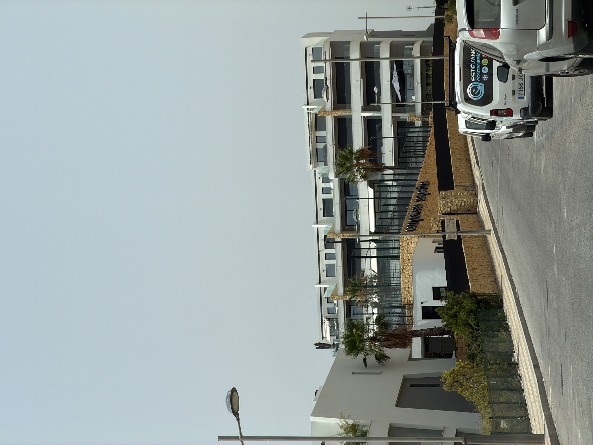 The image shows a street in Finestrat, Spain, with a modern apartment building in the background and several white vans parked on the right side of the road. The apartment building features a white and grey facade with multiple balconies and large windows. Palm trees are visible in front of the building. On the left side of the image, a white building with a sloped roof is partially visible. The road is asphalt, with a sidewalk lined with concrete and patterned tiles. The vans are parked in a line, with the rear of the closest van showing a logo with text that appears to say "ESTEVAN Fontaneria". The sky is clear and pale blue, suggesting a sunny day.