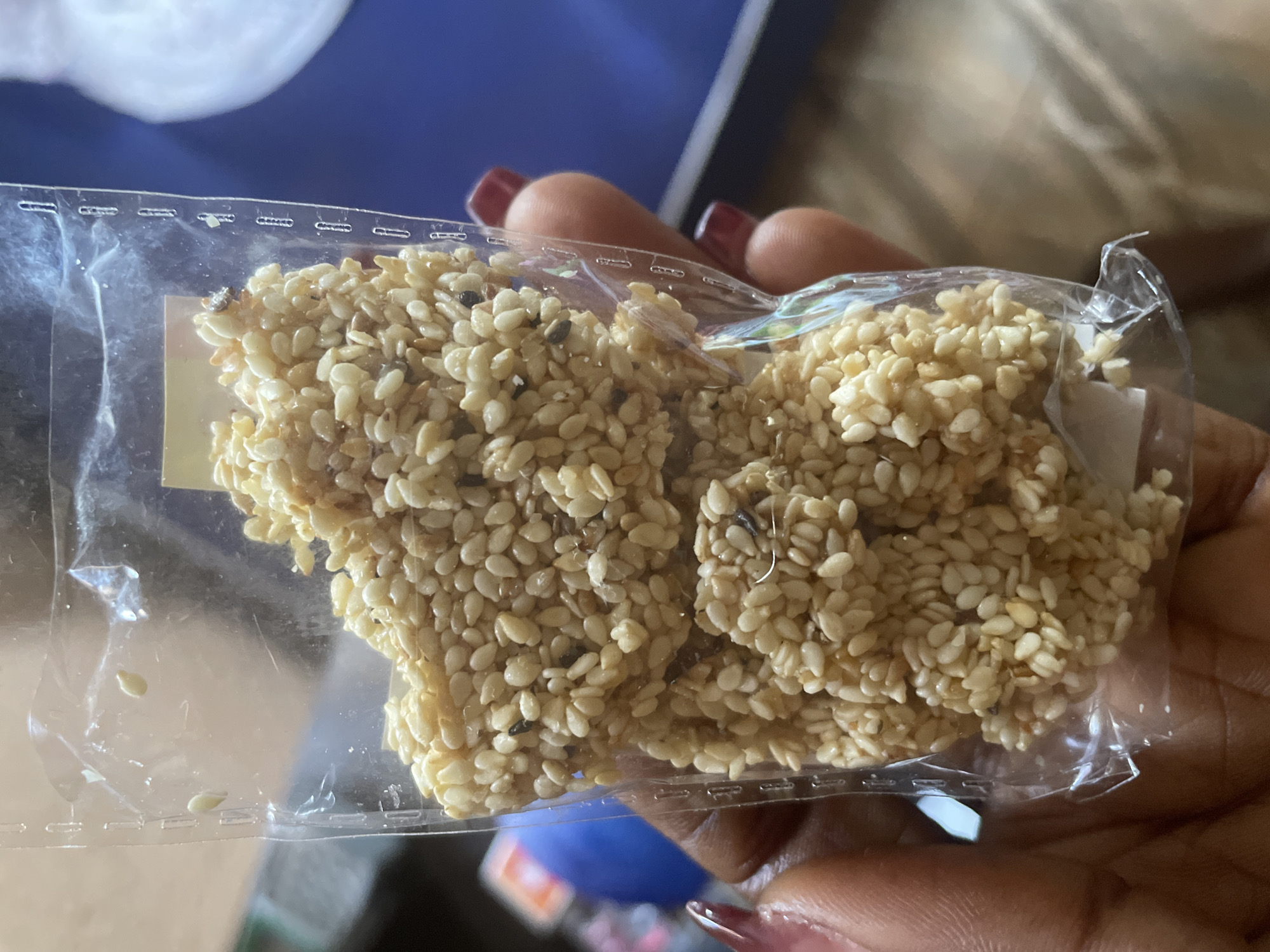 A close-up shot focuses on a person's hands holding a clear plastic package containing small, light-colored sesame seed candies. The candies appear to be slightly irregular in shape and are clustered together within the package. Some dark specks, possibly dirt or stray sesame seeds, are visible among the lighter ones. The person's fingernails are painted a dark red color. The background is blurred, showing hints of a blue surface and other indistinct objects, suggesting an indoor setting. There is no information in the image to indicate the location is Kakau, Nigeria.