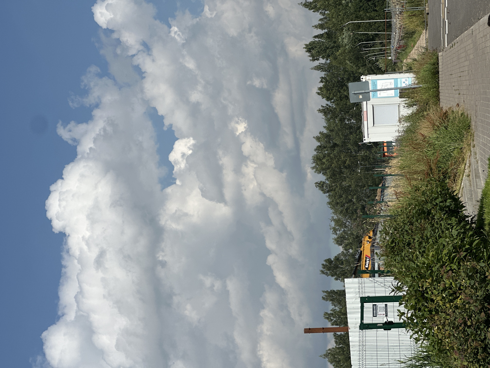 The image shows a construction site in Vsevolozhsk, Russia, under a partly cloudy blue sky. In the foreground, there is a paved walkway bordering a grassy area with dense green bushes. A white metal fence with a green gate encloses the site. A sign on the gate reads "ВНИМАНИЕ! ЧАСТНАЯ ТЕРРИТОРИЯ. ПРОХОД ЗАПРЕЩЁН." (ATTENTION! PRIVATE PROPERTY. ENTRY FORBIDDEN.) A tall, slender brown pole stands behind the fence.
Further into the site, a yellow SANY excavator is partially visible. A white portable building with a metal roll-up door and a blue informational sign is situated behind the bushes. A grey pole with a sign is placed in front of the portable building. Tall trees form a dense line across the background, with some power lines and lamp posts visible beyond them. The overall scene suggests an industrial or construction area.