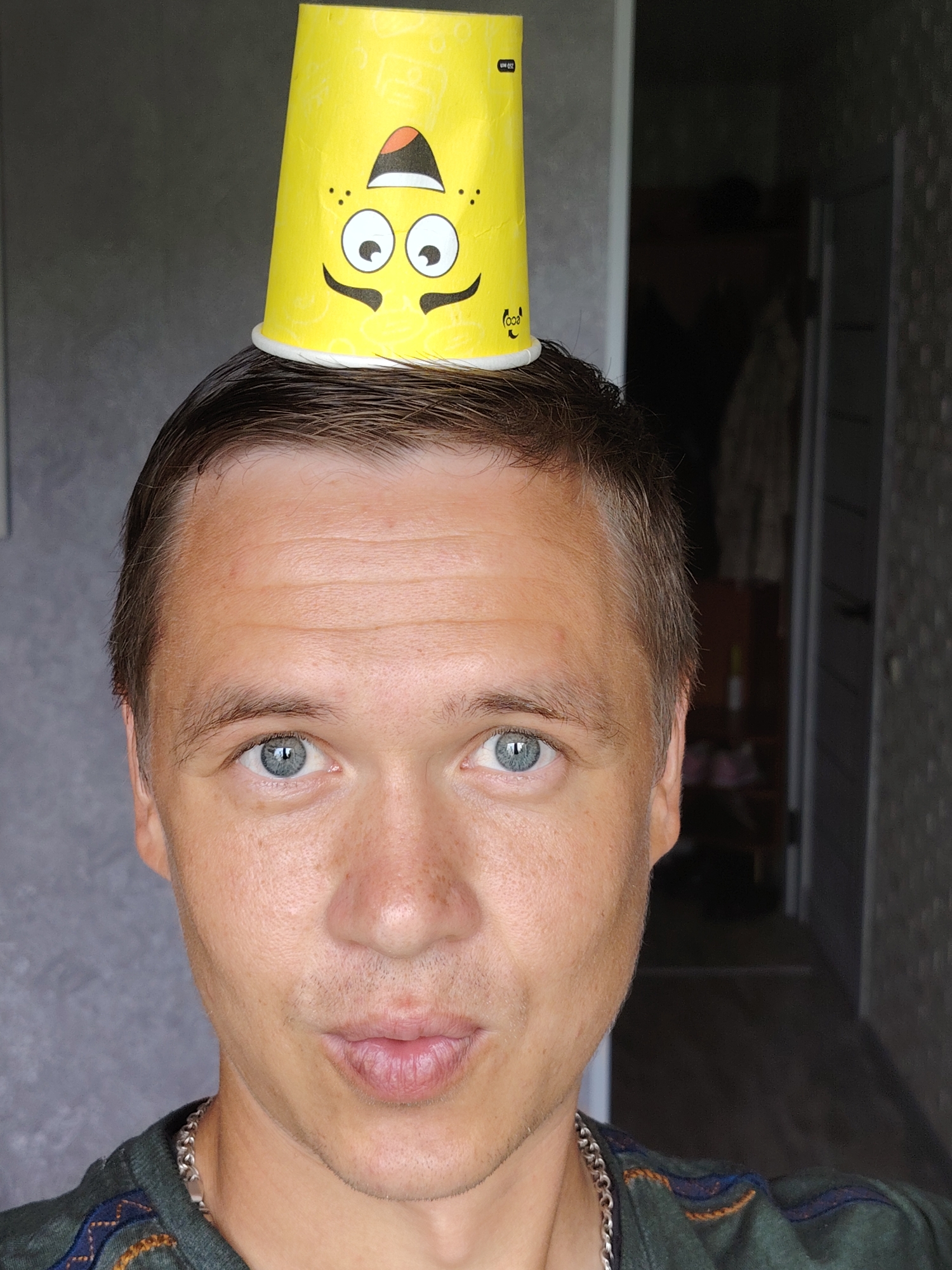 In Chaikovsky, Russia, a close-up, head-and-shoulders portrait shows a man with a yellow paper cup placed on his head. The cup features a cartoonish face with large eyes, a smiling mouth, and freckles. The man has light skin, short brown hair, and blue eyes. He is wearing a dark green shirt with an embroidered pattern around the collar and a silver chain necklace. The background is blurred, showing a wall and a doorway leading to a dimly lit interior space, consistent with a residential interior in Chaikovsky. The lighting is natural, coming from the front and left.