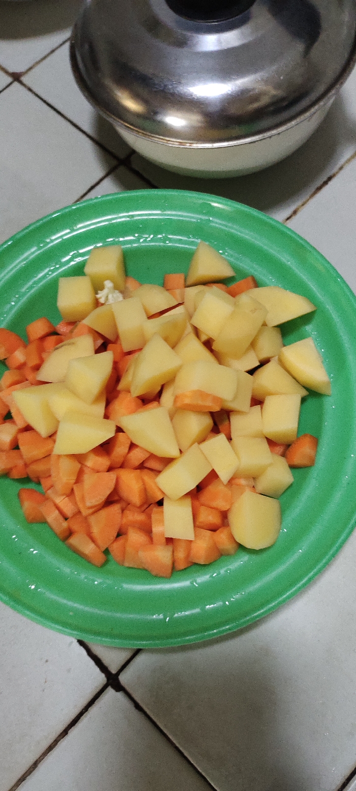 A green plastic plate filled with chopped potatoes and carrots sits on a tiled floor. The tiles are off-white and have dark grout lines. There is a silver pot lid out of focus in the background. The location is Kecamatan Lawang, Indonesia, based on the provided coordinates. The image suggests food preparation in a domestic kitchen setting. The potatoes and carrots are likely being prepared for a meal. The plate is the only subject of the image and is centered and in focus. The background is out of focus and shows only the floor. There is no sign of any people or other subjects in the image. The scene is static and depicts the food preparation process.