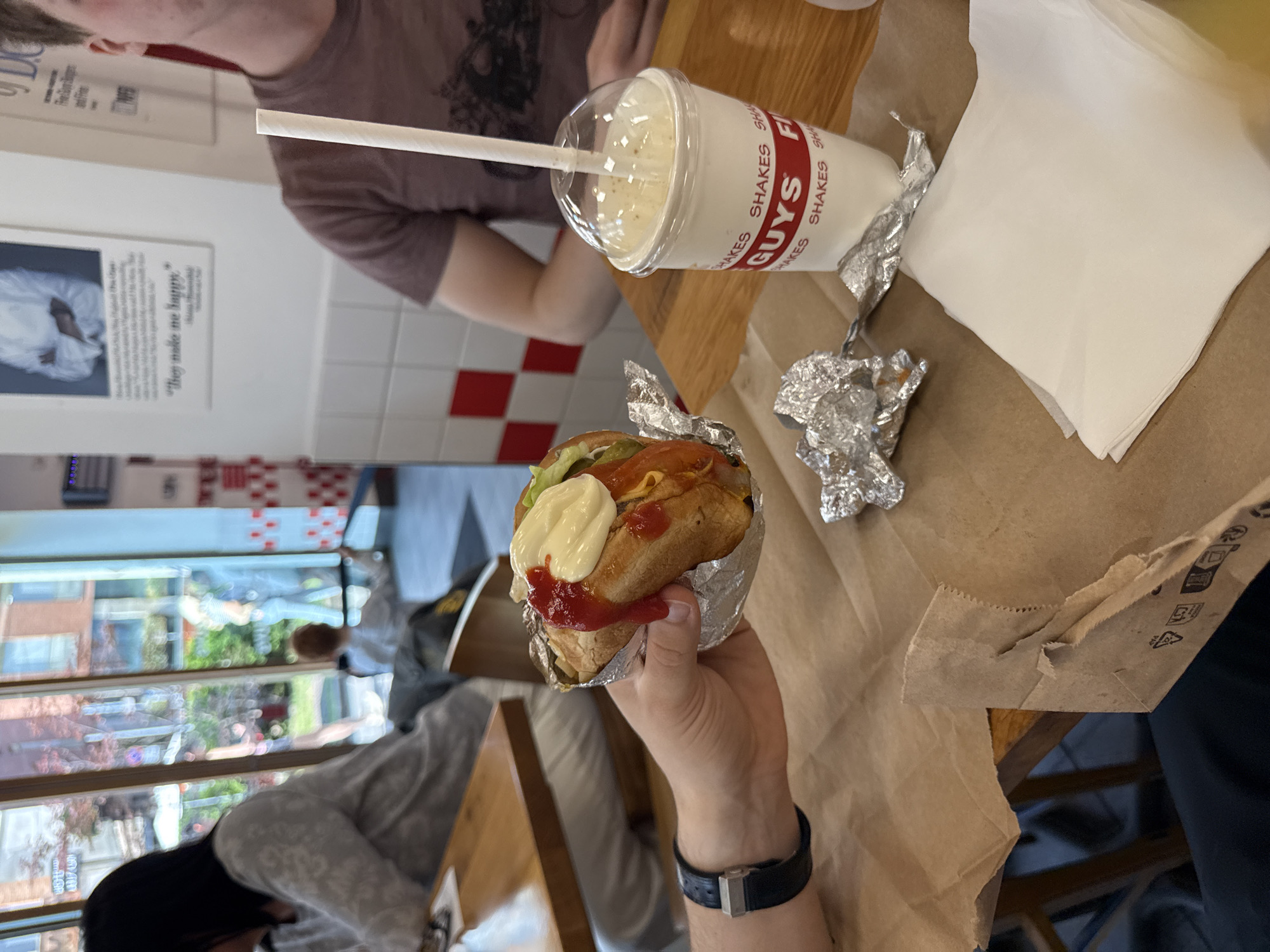 Here is a detailed description of the image:
**Scene:** An indoor dining setting, likely a restaurant. The viewpoint is from an individual seated at a wooden table. The table is covered with a brown paper bag and contains a Five Guys milkshake, a hamburger wrapped in foil, crumpled foil, and napkins. A person is holding the hamburger in their left hand, and part of their arm and wrist is visible. A black watch is on their wrist. Another person is partially visible in the background, seated on the same side of the table.
**Objects:**
* **Food:** A hamburger with toppings including lettuce, tomato, ketchup, and mayonnaise.
* **Drink:** A Five Guys milkshake with a white straw.
* **Tableware:** Crumpled foil, napkins.
* **Restaurant décor:** A red and white tiled wall can be seen in the background. There is a framed print on the wall with the text '"They make me happy."'
* **Clothing and Accessories:** A black watch with a silver clasp. The person in the background is wearing a gray-brown shirt.
**Environment:** Natural light is streaming in through a large window that shows an exterior street scene with foliage and buildings.
**People:** Two individuals are partially visible. One is holding the hamburger; the other is seated in the background.
**Actions/Interactions:** One person is holding a hamburger, likely preparing to eat it. It is implied that the people are eating at a restaurant.
**Location Information:** The restaurant appears to be a Five Guys location.