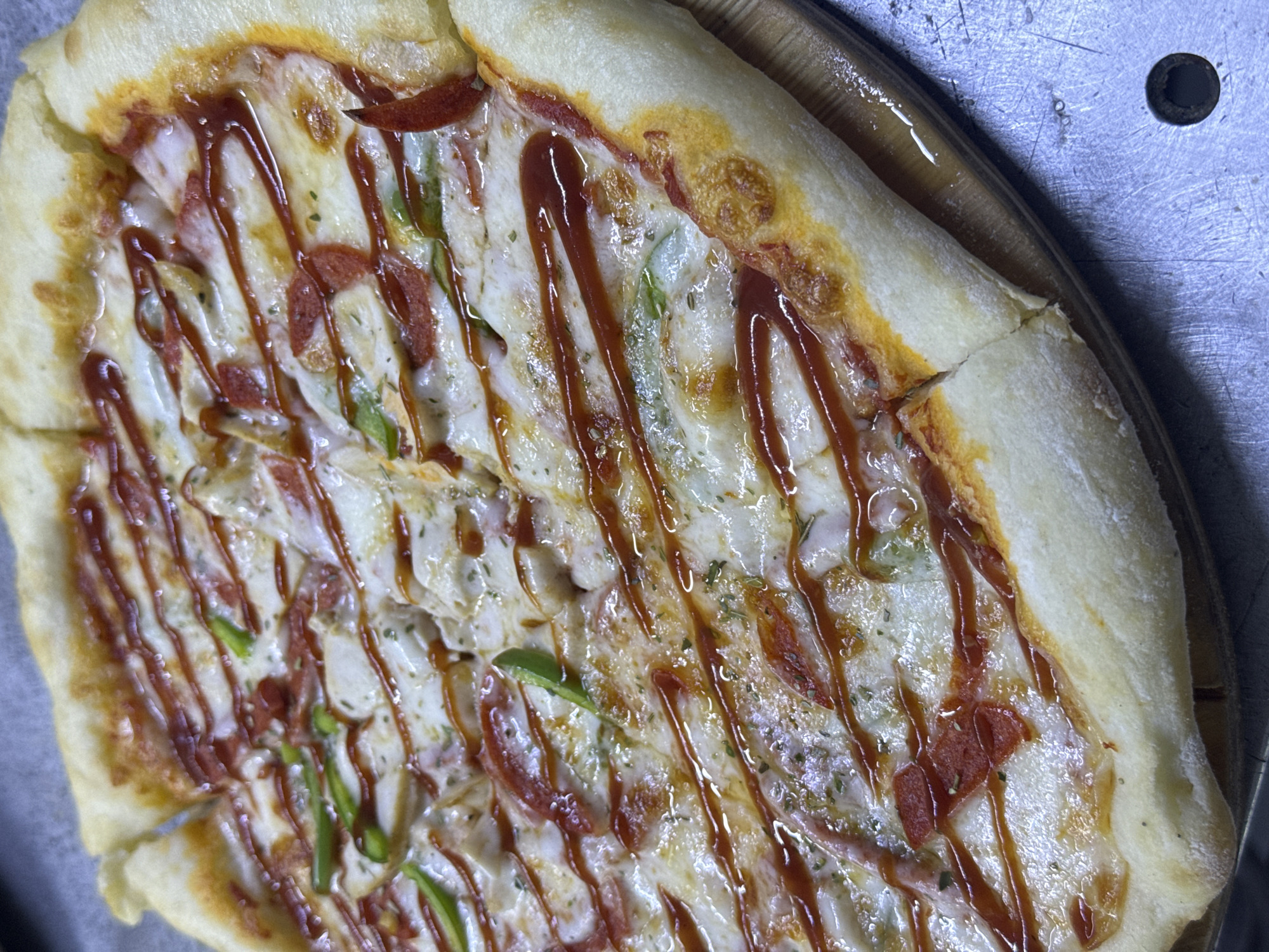 A close-up, high-angle shot shows a freshly made pizza with a thick, golden-brown crust. The pizza is sliced into multiple pieces and topped with melted cheese, pepperoni slices, green peppers, and a drizzle of a dark red sauce, possibly barbecue sauce or ketchup. The crust has a slightly puffed and airy texture, with some darker browned spots indicating it was baked to perfection. The pizza rests on a wooden pizza peel, which is placed on a textured metal surface. The lighting is dim and creates subtle shadows, highlighting the glossy sheen of the sauce and cheese. The setting appears to be a commercial kitchen or a restaurant environment in the city of Ақши, Kazakhstan, possibly during a busy service. There are no people visible in the frame.