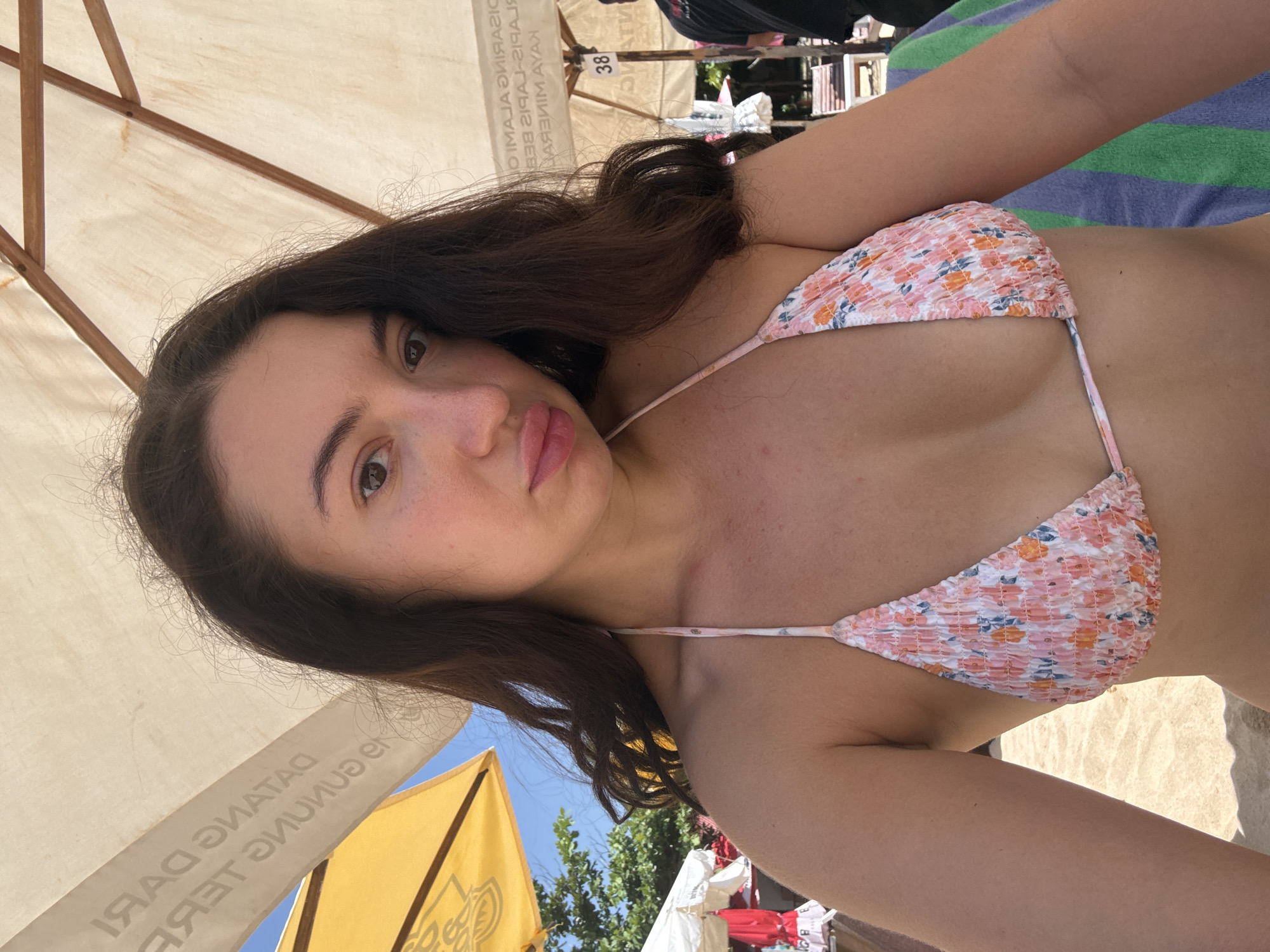 A young female with long, dark brown hair and light skin, wearing a pink floral-patterned and ruched bikini top, is depicted in a close-up, selfie-style photograph. She looks directly into the camera with slightly pursed lips. She is seated or reclined under a large, light-colored sun umbrella. Mirrored text is visible on the underside of this umbrella, including partial phrases such as "DATANG GARUDA" and "KAYA MINERAL," indicative of the Indonesian language. To the left, a bright yellow umbrella is visible against a clear blue sky and tropical foliage. Further in the background, other white umbrellas and wooden beach chairs are discernible, along with a partial view of another individual in a dark shirt. A sign displaying the number "38" is attached to a wooden support beam of an umbrella frame on the right. A multi-striped towel (blue, green) is partially visible at the bottom right of the frame. The ground beneath appears to be sand. The scene is located on a beach in Nusa Dua, Indonesia.