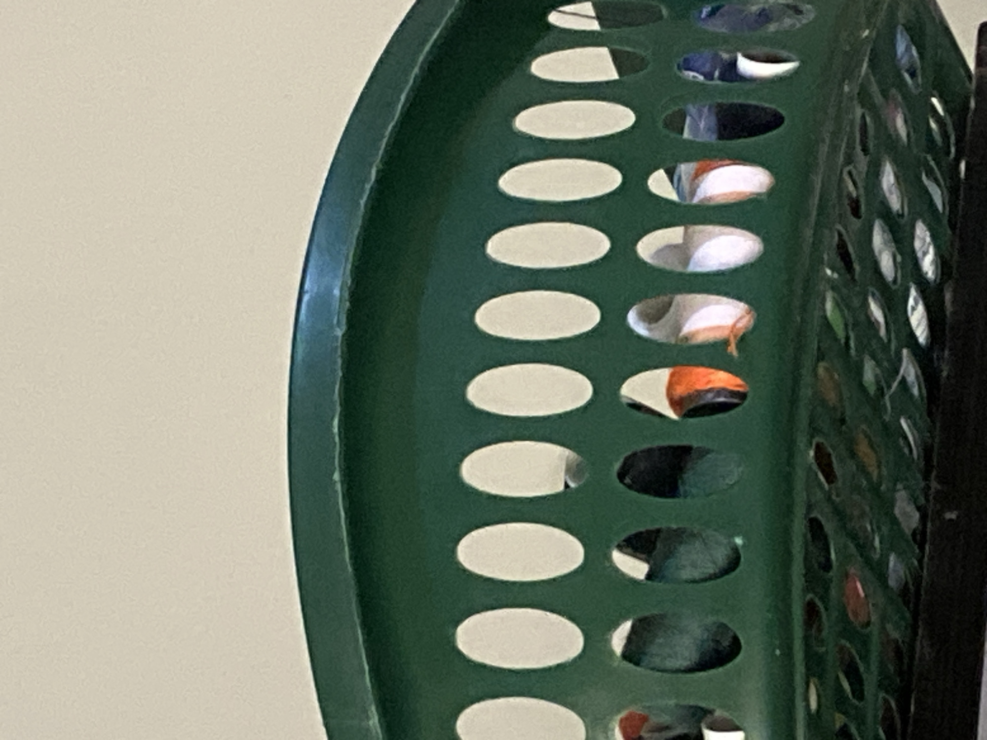The image shows a close-up view of the top rim and upper portion of a green plastic laundry basket filled with clothing. The basket has a pattern of oval-shaped holes along its sides. Through these holes, various articles of clothing are visible, including items with orange and white colors, as well as darker fabrics. The basket is positioned on a dark surface, likely a shelf or the floor, with a light-colored wall in the background. There are no people or specific landmarks visible that would indicate a location in Jauro Yinu, Nigeria. The scene appears to be indoors, likely in a domestic setting.