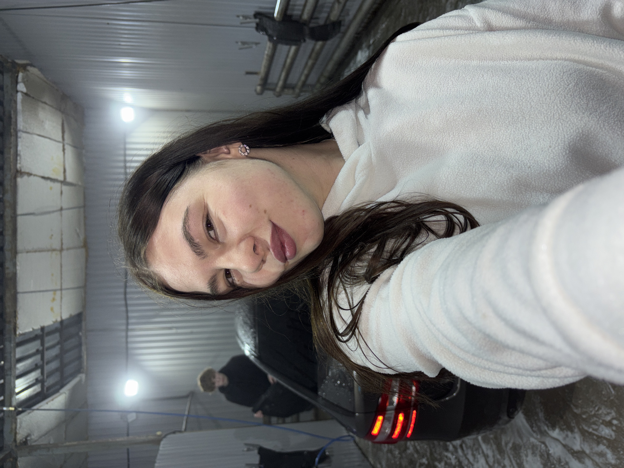 A young woman is captured in a close-up selfie taken indoors, likely at a car wash or industrial garage in Chaikovsky, Russia. She is positioned prominently in the foreground, with her head tilted, looking towards the camera with a subtle smile. Her long, dark hair cascades over her shoulders and back. She is wearing a white, fuzzy hooded top, possibly fleece, and a small, circular, jeweled earring is visible in her left ear.
In the mid-ground, the rear section of a dark-colored car is visible, featuring illuminated red taillights. A blue hose or cable is on the wet ground near the vehicle. The background reveals two blurry figures; one appears to be standing or bending near the car, suggesting activity related to vehicle care. The setting features concrete block or tiled walls, some displaying wear, and a corrugated metal ceiling. Bright artificial lights overhead create reflections and a slight haze in the atmosphere. The ground is wet, consistent with a car washing environment. No visible text is present in the image. The indoor setting and artificial lighting do not indicate a specific time of day.