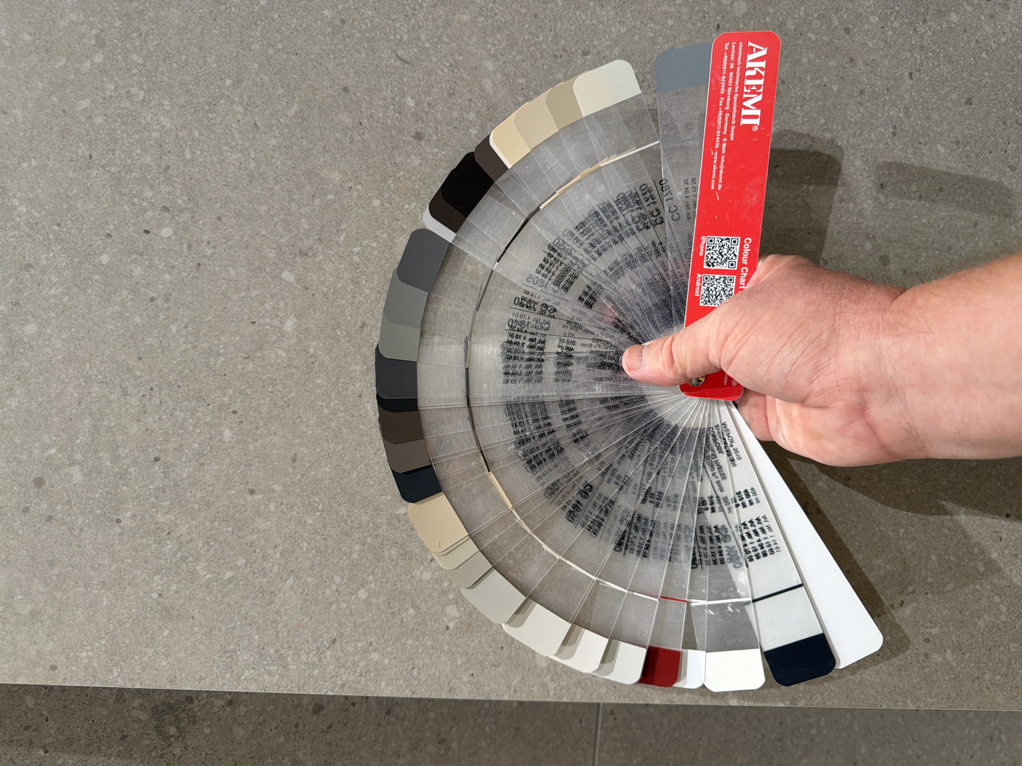 An overhead medium shot shows a hand holding a fan deck of paint color swatches against a textured, neutral-toned floor. The paint swatches are arranged in a semi-circle and range in color from white, cream, beige, brown, gray, to black. Some of the swatches are transparent with printed text and QR codes. The floor appears to be a large format tile with a slightly speckled pattern. The scene is brightly lit, and the background is out of focus. There is no identifiable context for the location within the city of Jerusalem, Israel.