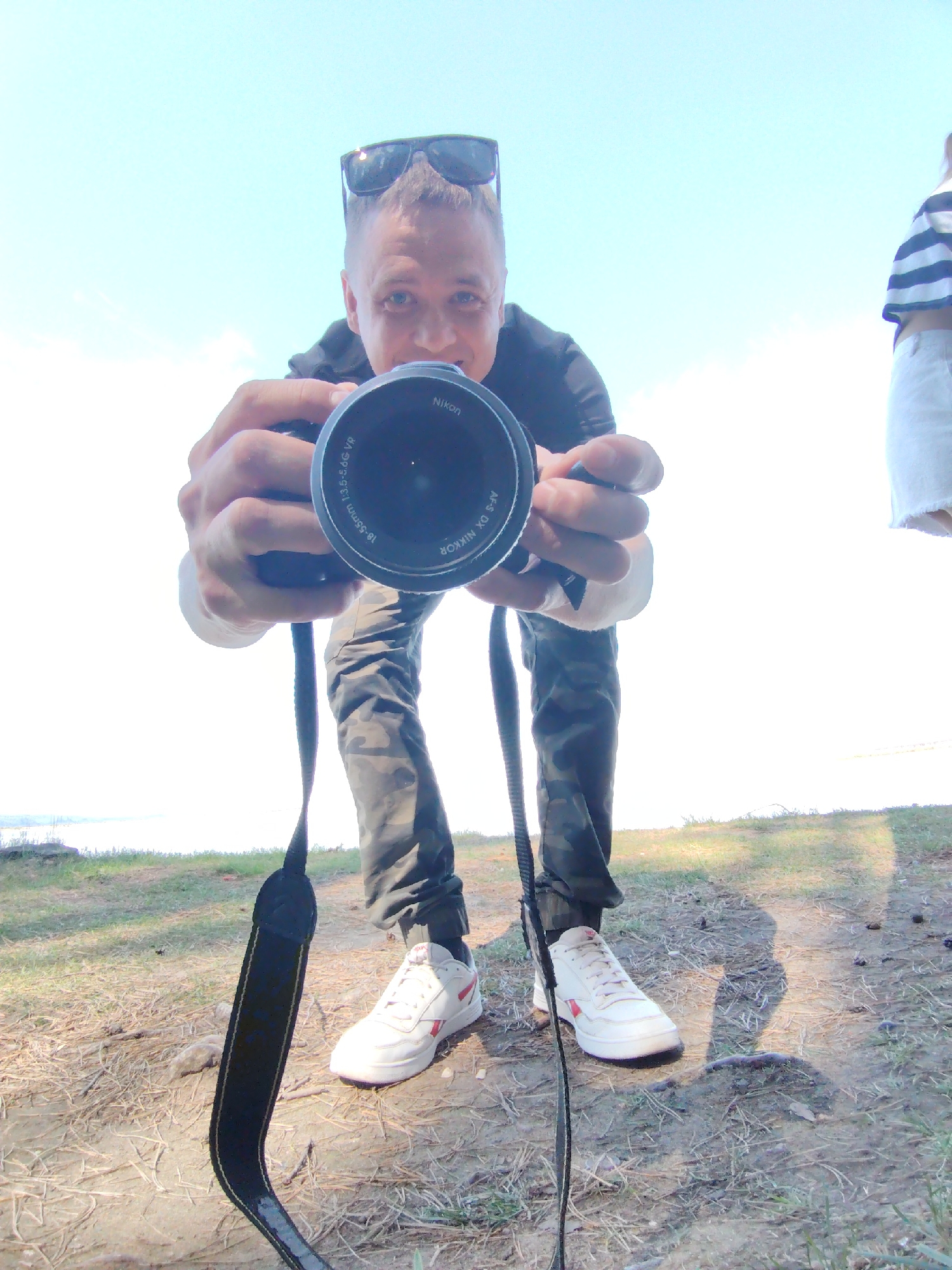 A man stands outdoors holding a black Nikon camera with an 18-55mm lens. The man is wearing a black t-shirt, camouflage pants, and white sneakers with red accents. He has sunglasses perched on top of his head. The camera strap is visible around his neck. In the background, there is a blurry figure of a person in a striped shirt and white pants, grass, and the edge of what appears to be a body of water. The location may be in the city of Chaikovsky, Russia, but it is not explicitly discernable from the image.