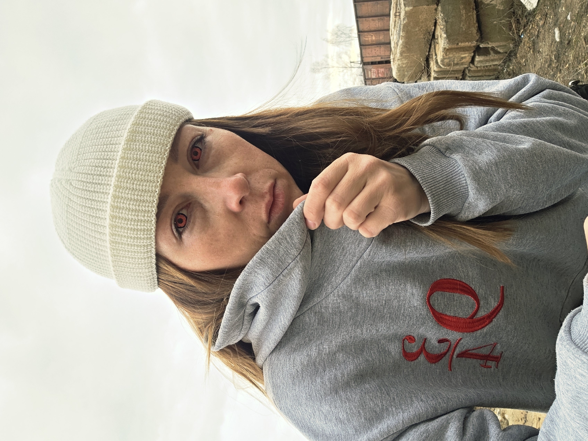 A close-up portrait features a woman with long, reddish-brown hair wearing a white knit beanie and a grey hoodie. Her eyes are a striking feature, displaying bright red irises, possibly from colored contact lenses. She looks directly at the camera with a neutral to slightly serious expression. Her right hand is visible, pulling up the collar of her hoodie. The grey hoodie has red embroidered text on the chest, clearly showing "9 3/4".
The setting is outdoors under an overcast or bright sky, which dominates the upper portion of the background. To the viewer's right, in the blurry distance, are bare trees and what appears to be an old, reddish-brown structure, possibly industrial or part of a train car. To the viewer's left, there are stacked, rough-hewn stone or concrete blocks. The overall lighting is soft and diffused, indicating an overcast day.