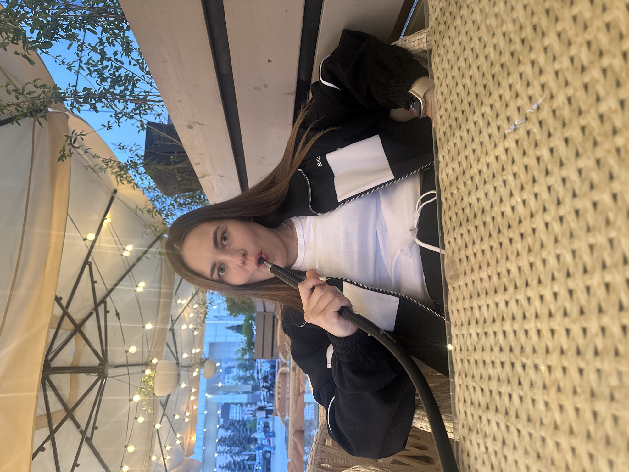 A young woman with long brown hair is sitting outdoors at a table with a woven surface, holding a hookah hose to her mouth. She is wearing a black jacket with white accents and a white t-shirt. A smartwatch is visible on her left wrist. In the background, there is a light-colored awning with string lights, and beyond that, a cityscape with buildings and trees under a blue sky. The scene appears to be in an outdoor seating area, possibly a cafe or restaurant. Given the text on the jacket, the location is likely Perm, Russia.